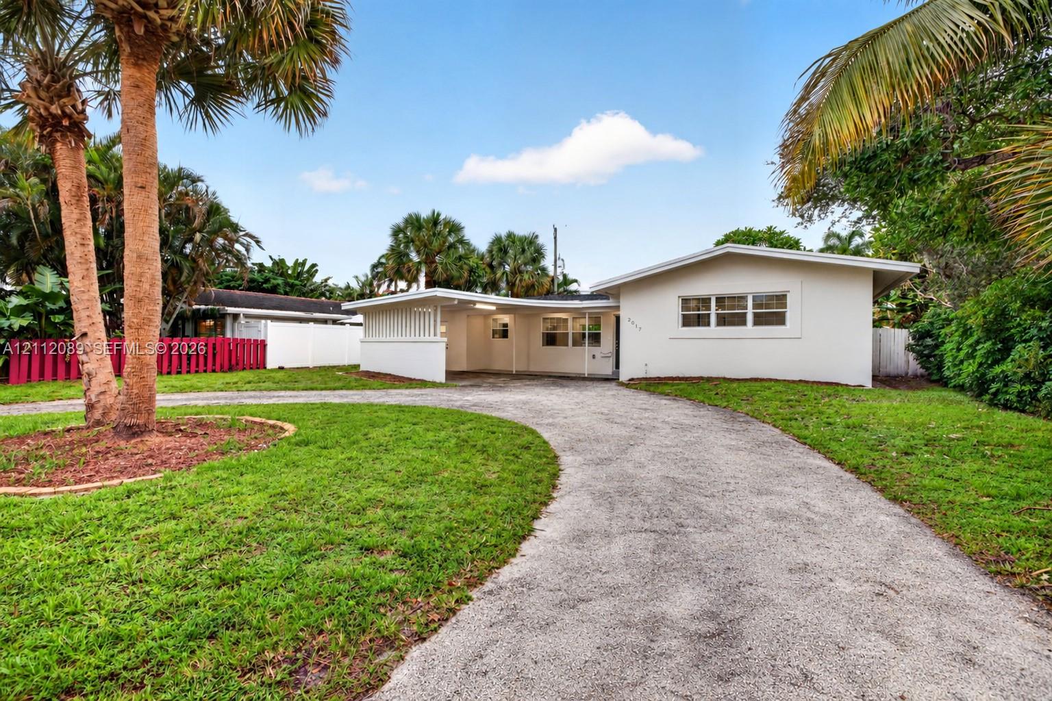 Single-story exterior featuring a light-colored facade, attached carport with slatted screening, and a curved paved driveway set within a manicured landscape