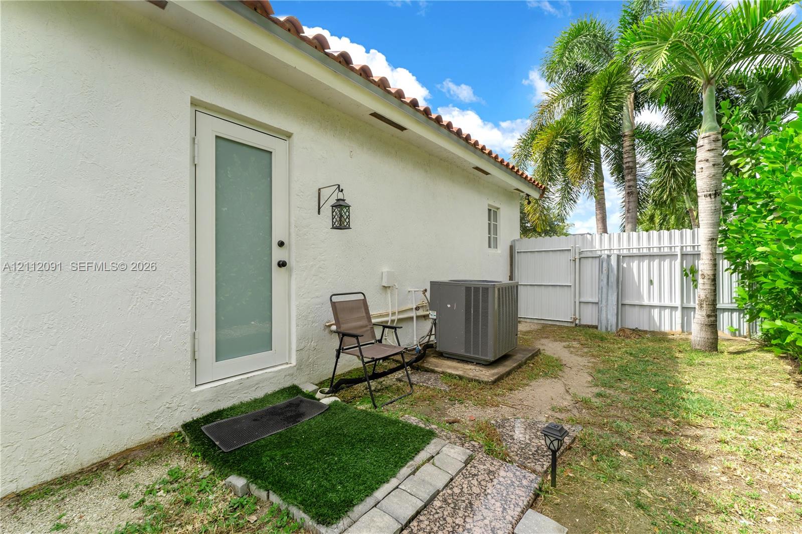 Exterior side yard featuring white stucco siding, a glass-panel door, terracotta roof tiles, a white vinyl privacy fence, and mature palm trees
