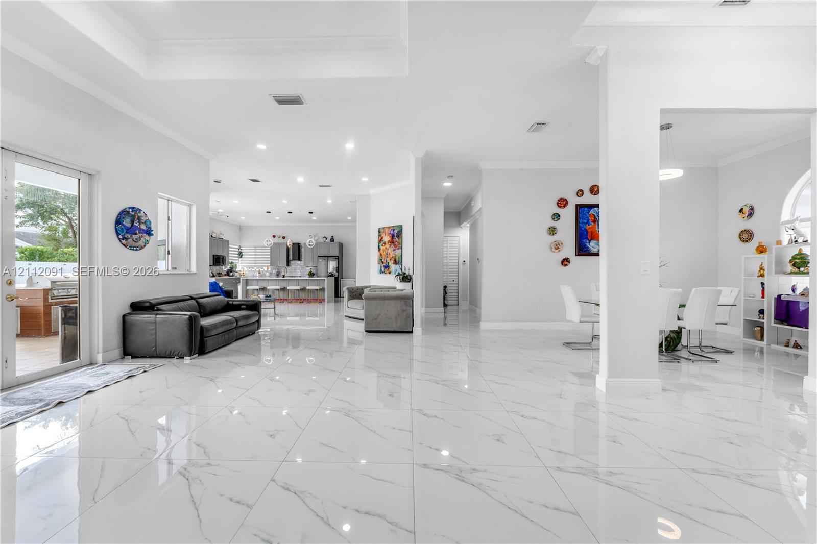 Open-concept main living area featuring large-format polished tile flooring, recessed lighting, tray ceilings, and a glass-paneled door leading to an outdoor space