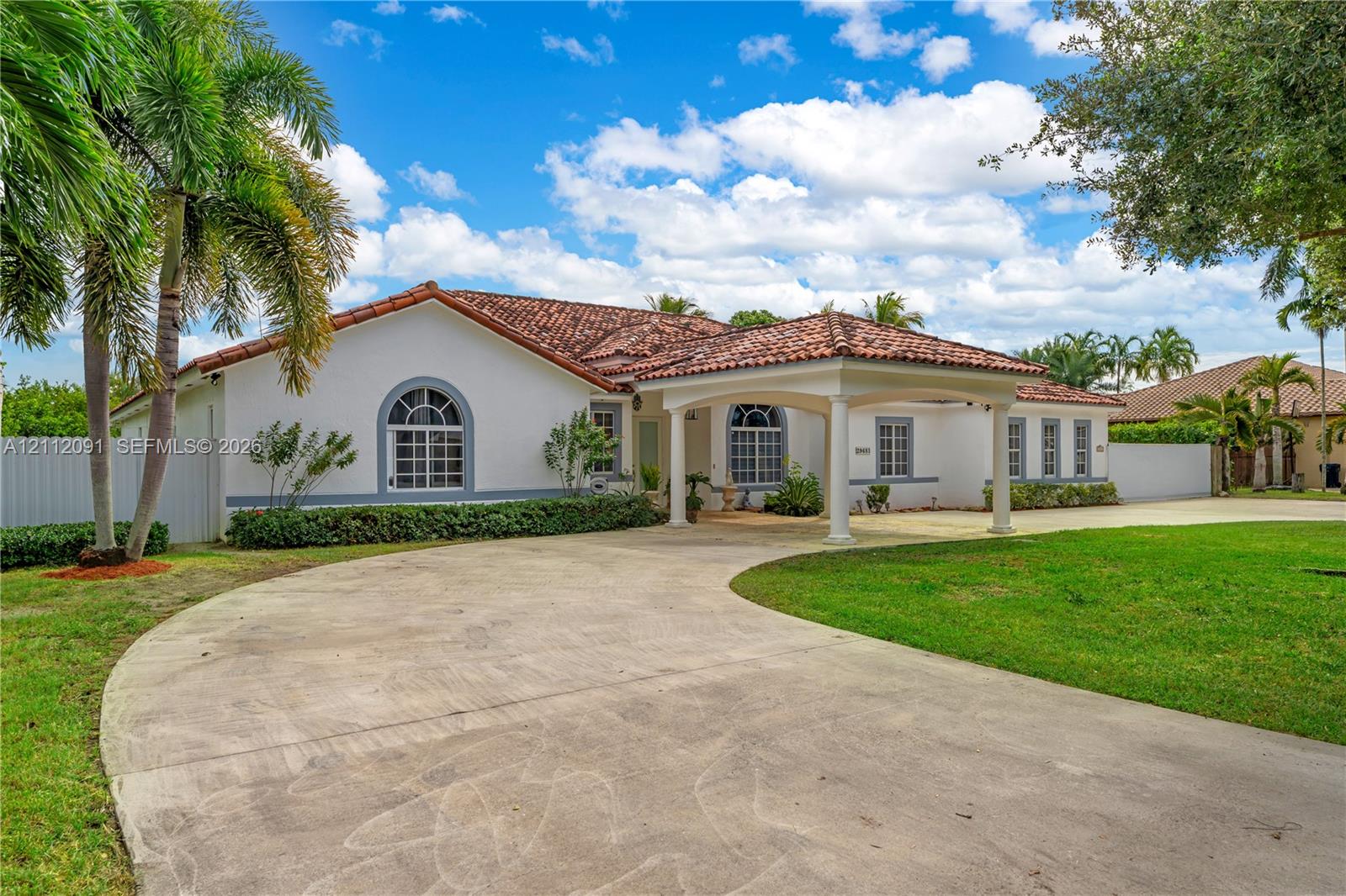 Single-story residence featuring a tile roof, arched windows, a covered porte-cochre, and a circular concrete driveway set on a landscaped lot Single-story residence featuring a tile roof, arched windows, a covered porte-cochre, and a circular concrete driveway set on a landscaped lot