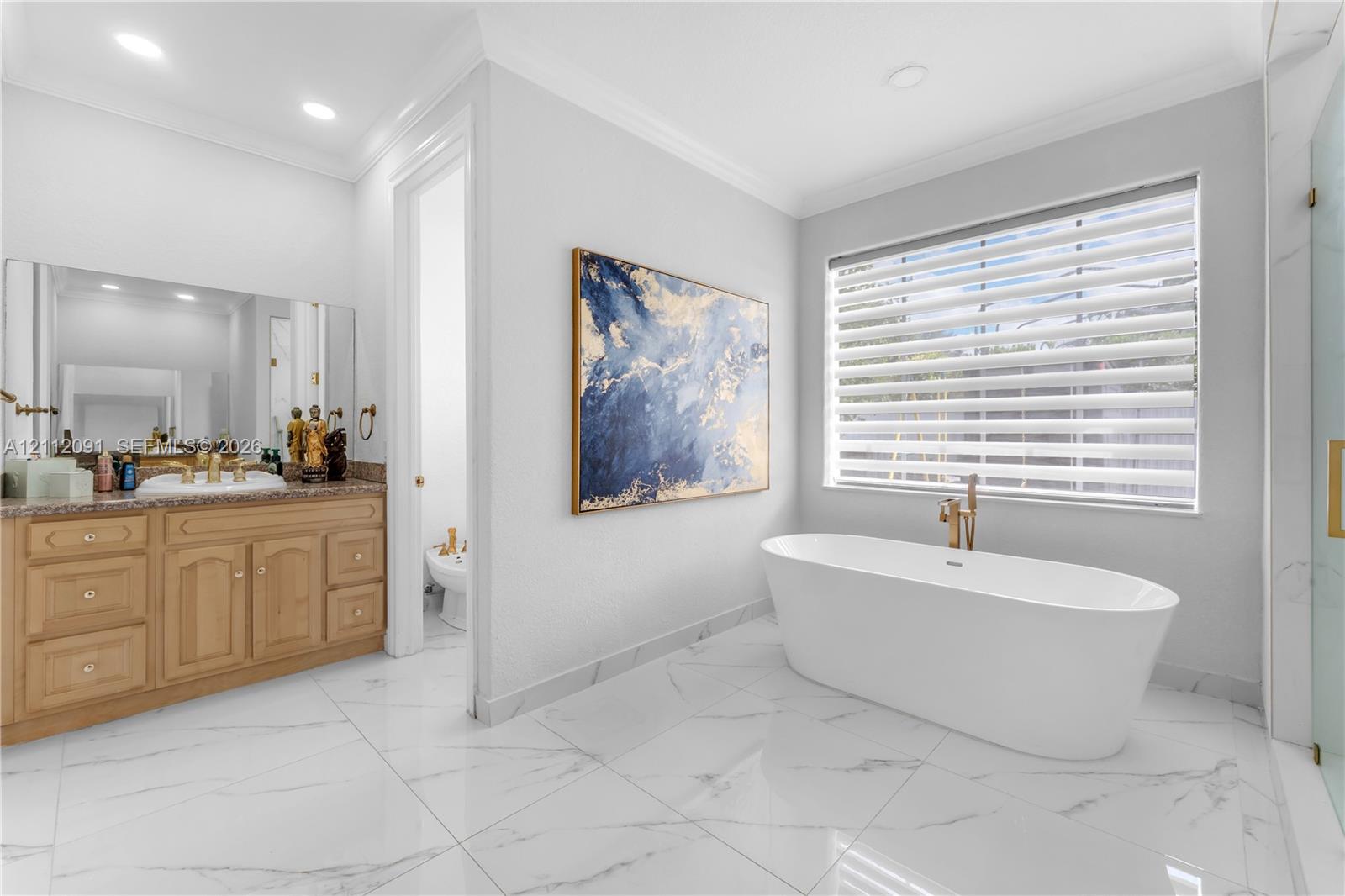 Freestanding soaking tub with gold-tone fixtures, wood cabinetry with stone-surface countertop, white tile flooring with veining detail, and large-format wall mirror