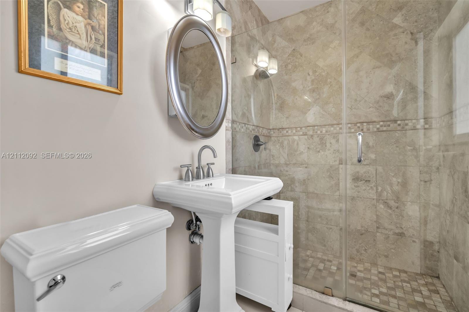 Bathroom featuring a pedestal sink, oval wall-mounted mirror, glass-enclosed shower with stone-tile surround, and mosaic tile shower flooring