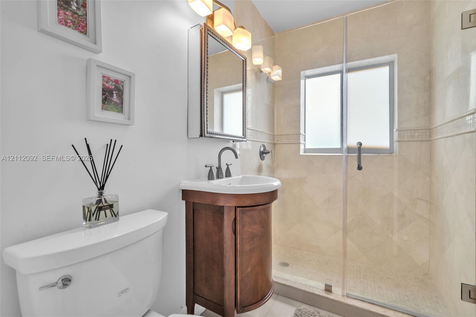 Curved wood-finish vanity with a white basin, stone-tiled shower with a glass enclosure, and wall-mounted three-light fixture