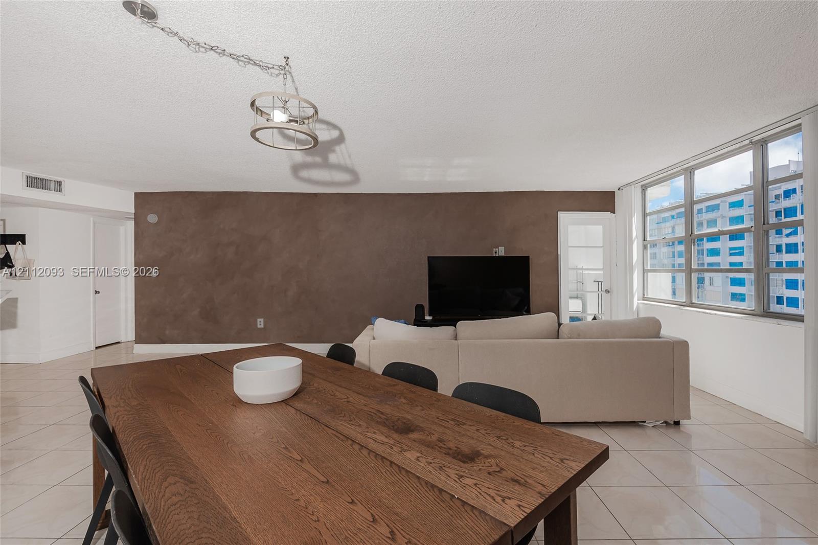 Open-concept living space featuring large-format ceramic tile flooring, a prominent accent wall with textured finish, expansive multi-pane windows, and a suspended industrial-style drum pendant light fixture