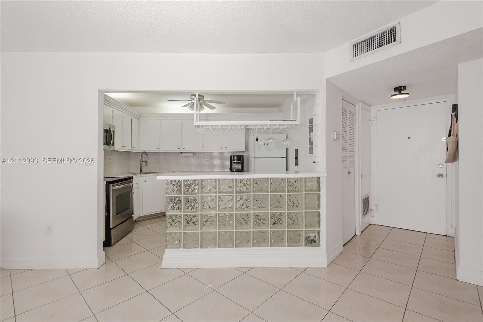 Kitchen featuring white cabinetry, stainless steel appliances, a glass-block breakfast bar, and a hanging stemware rack over ceramic tile flooring Kitchen featuring white cabinetry, stainless steel appliances, a glass-block breakfast bar, and a hanging stemware rack over ceramic tile flooring