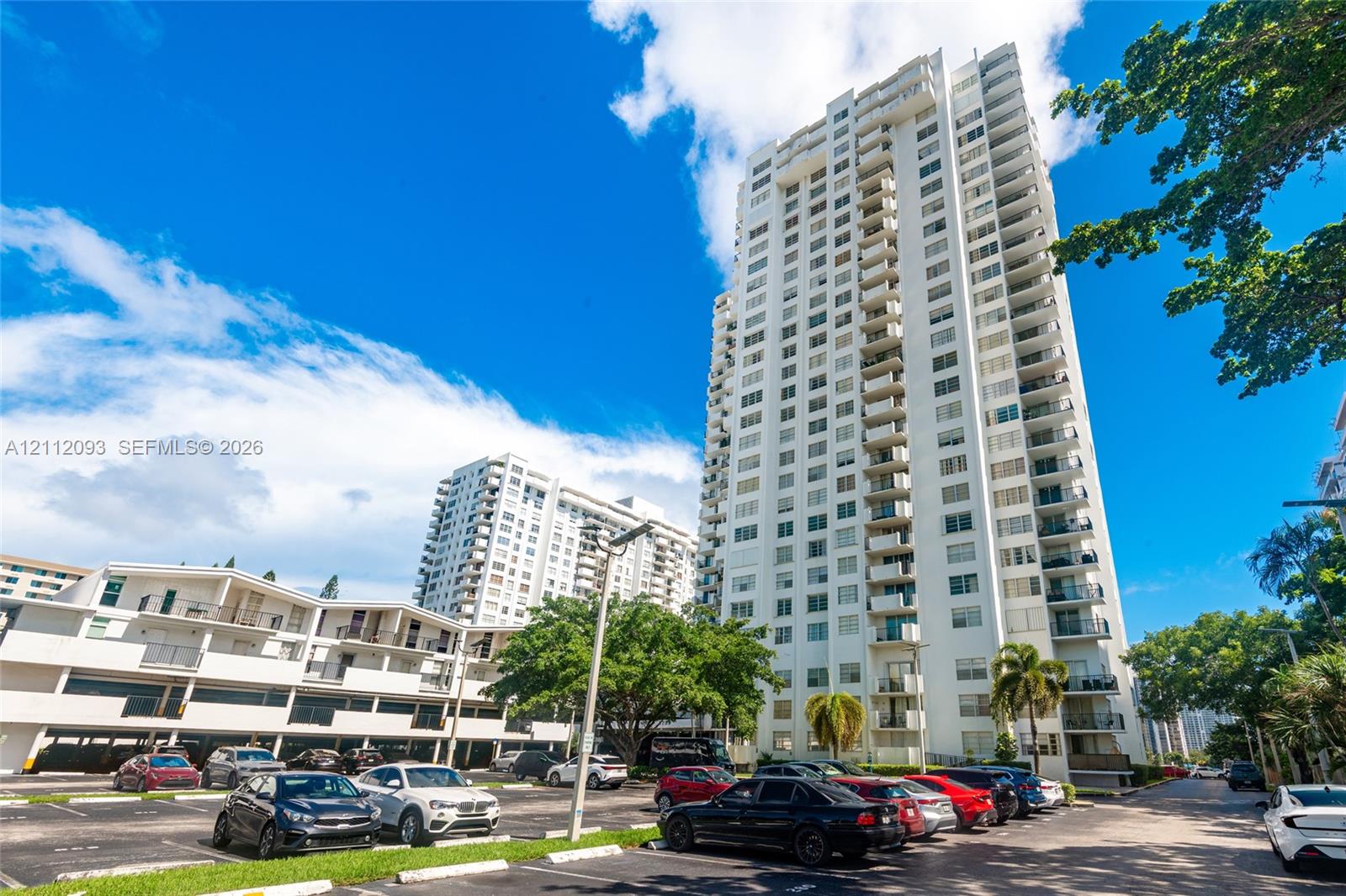 High-rise residential tower featuring white stucco exterior, multiple stacked balconies, and paved surface parking