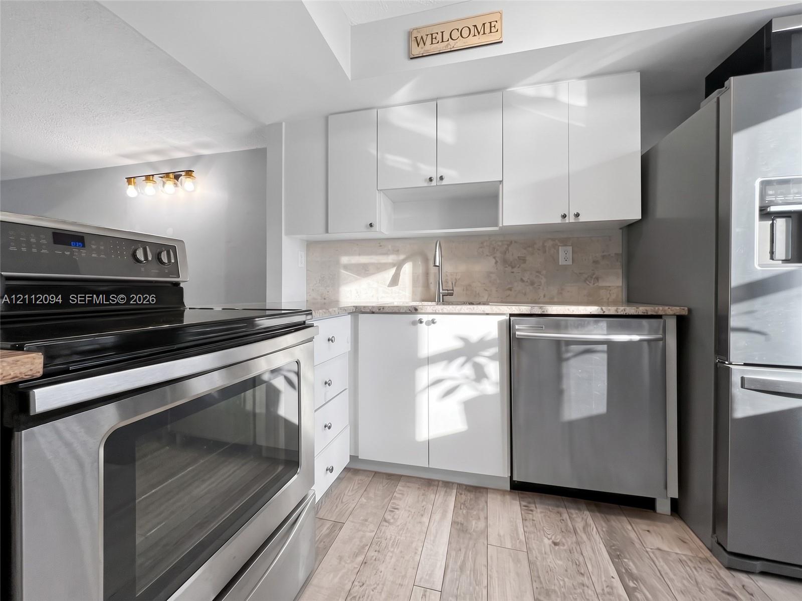 Kitchen featuring white flat-panel cabinetry, stainless steel appliances, neutral stone-finish backsplash, and wood-finish flooring Kitchen featuring white flat-panel cabinetry, stainless steel appliances, neutral stone-finish backsplash, and wood-finish flooring