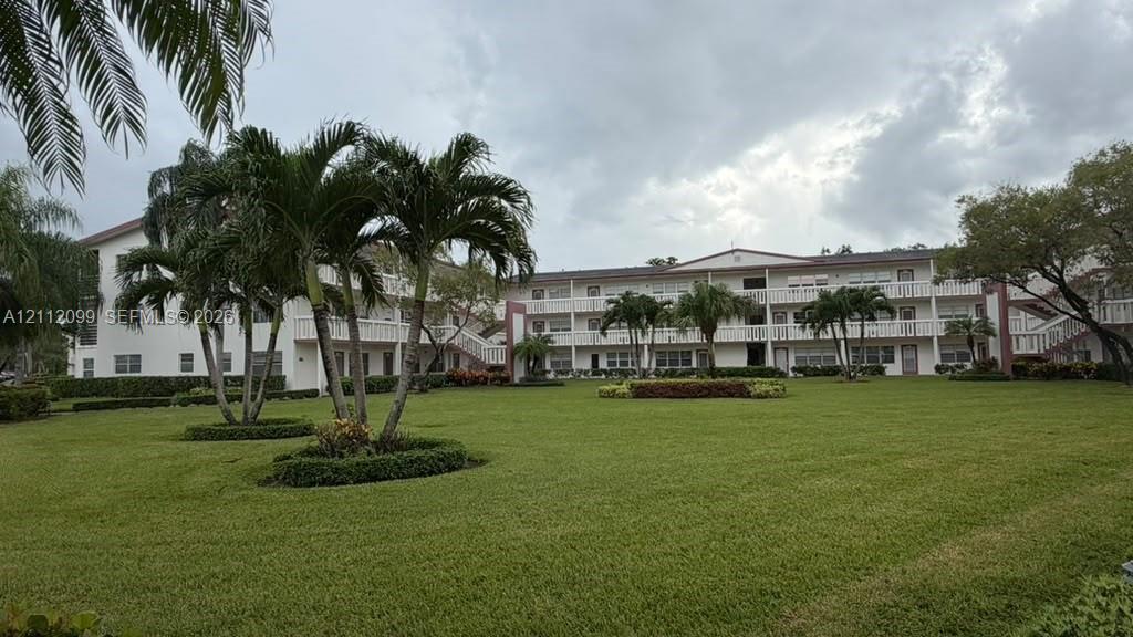 Multi-level exterior featuring a light-toned stucco faade, open-air balcony railings, expansive manicured lawn, and mature palm tree landscaping