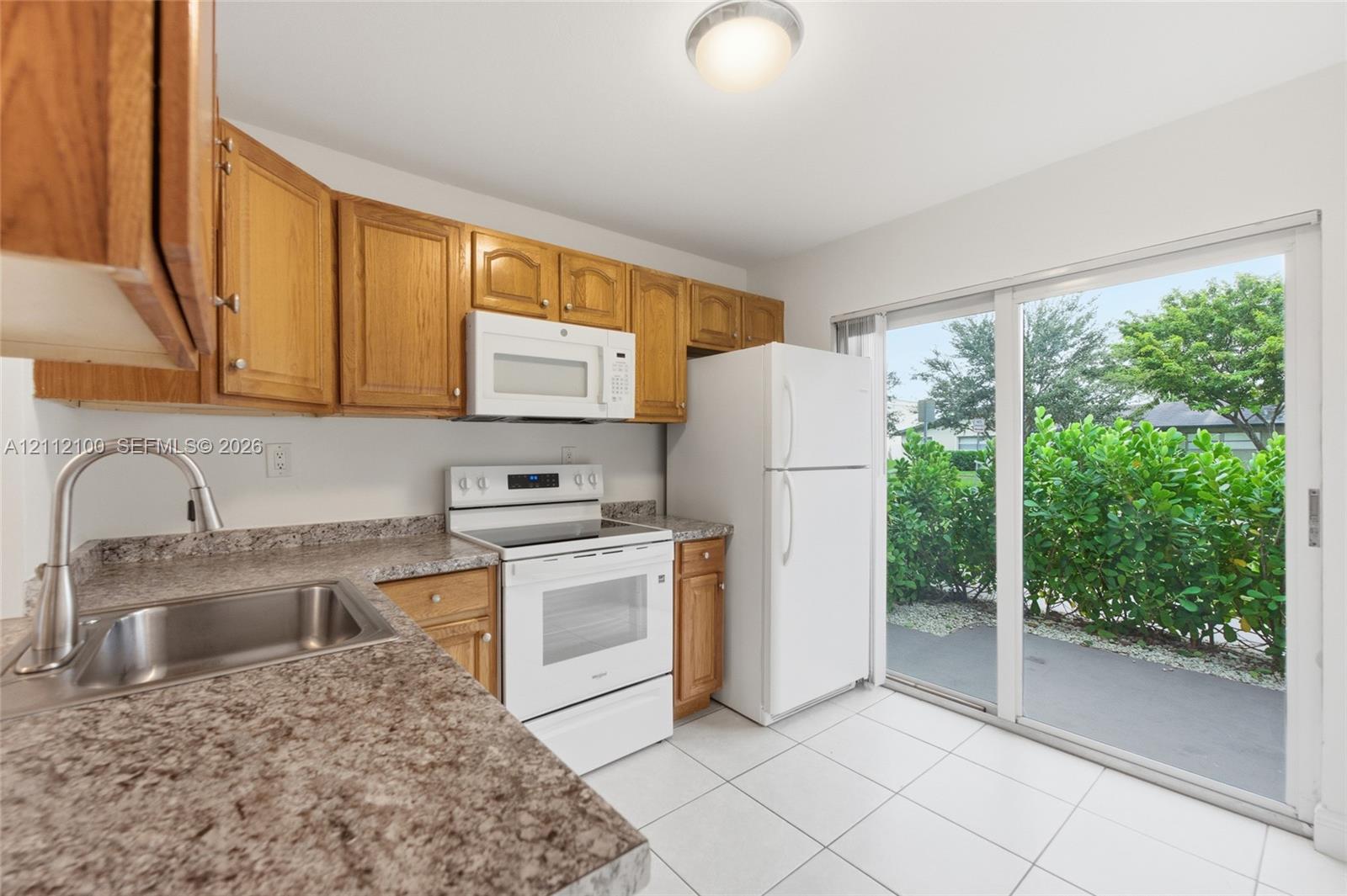 Kitchen featuring wood cabinetry, stone-finish countertops, stainless steel sink with high-arch faucet, white appliances, and a sliding glass door