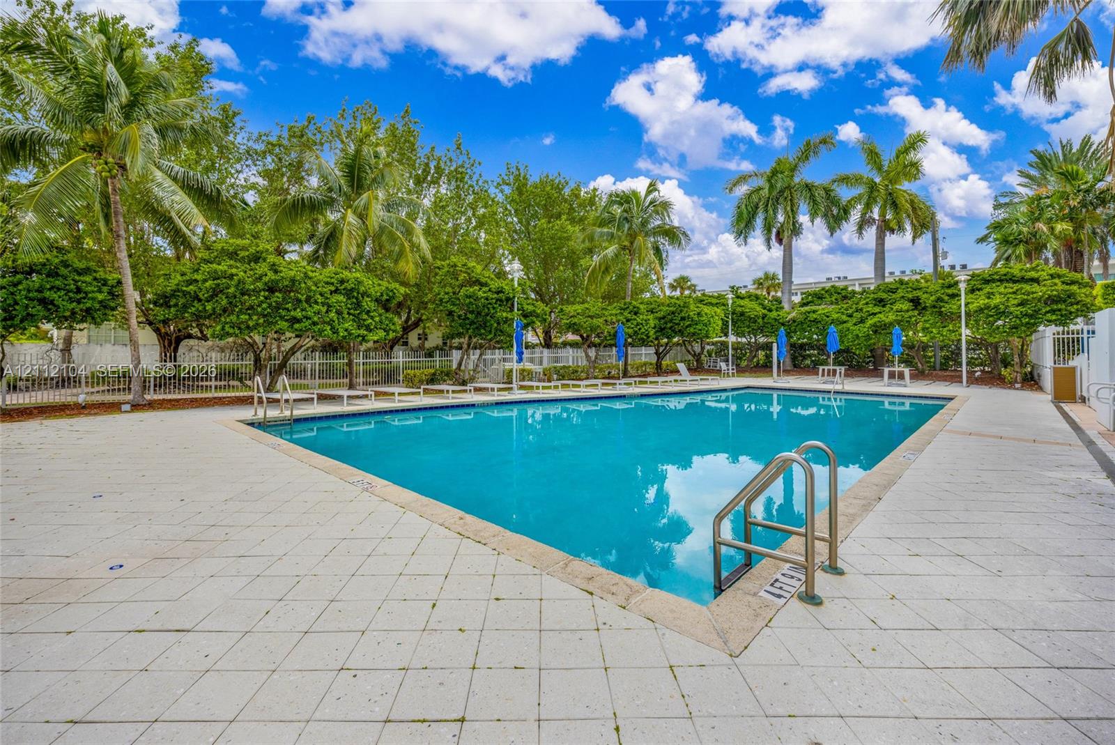 Swimming pool with light-toned tiled deck, metal handrails, and surrounding perimeter fencing set amidst mature landscaping and palm trees