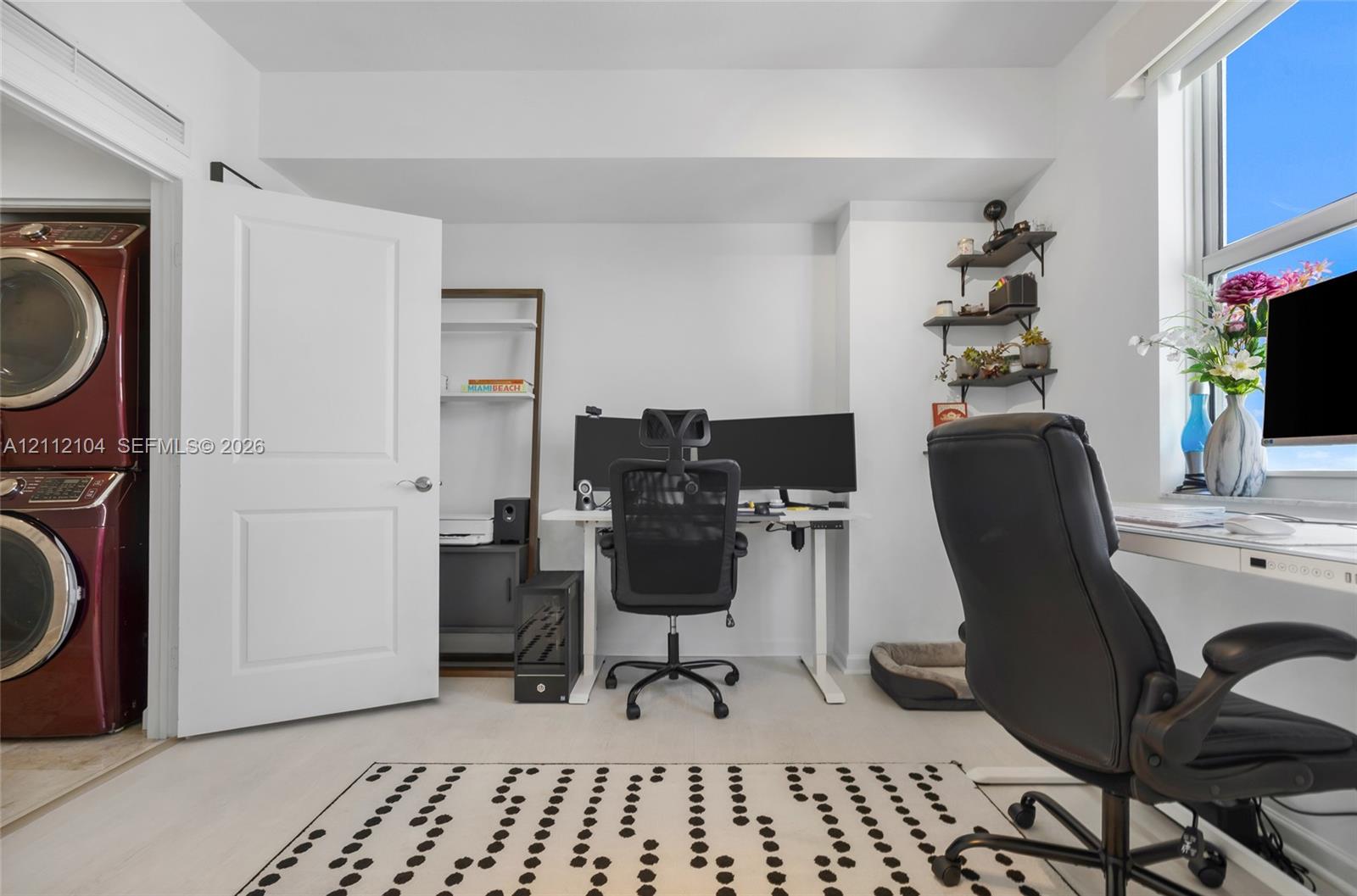 Room featuring neutral wall-to-wall carpeting, a large window, wall-mounted tiered shelving, and an enclosed laundry closet with stacked washer and dryer unit