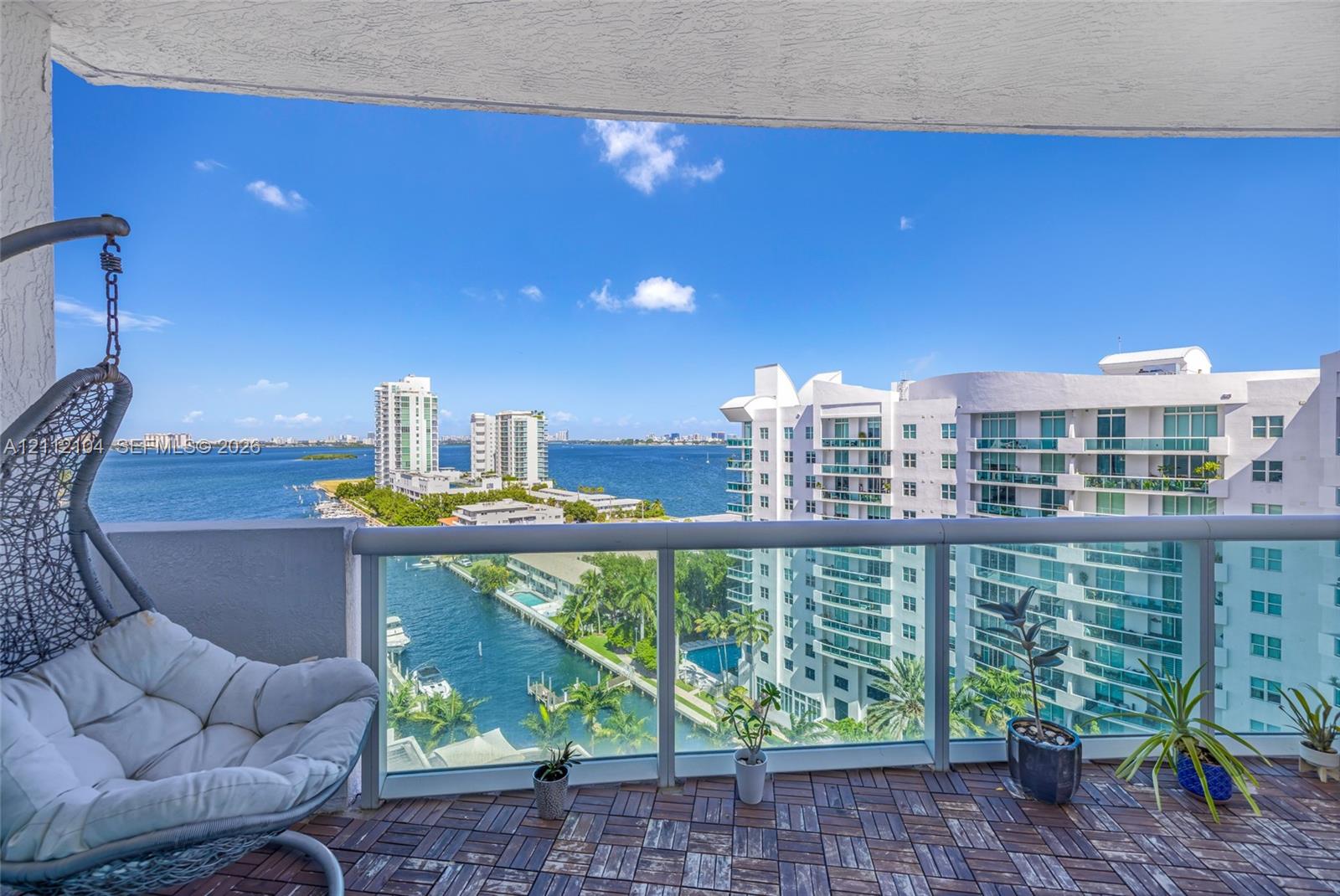 Covered balcony featuring wood-finish deck tiling and a glass-panel railing, overlooking water and urban scenery