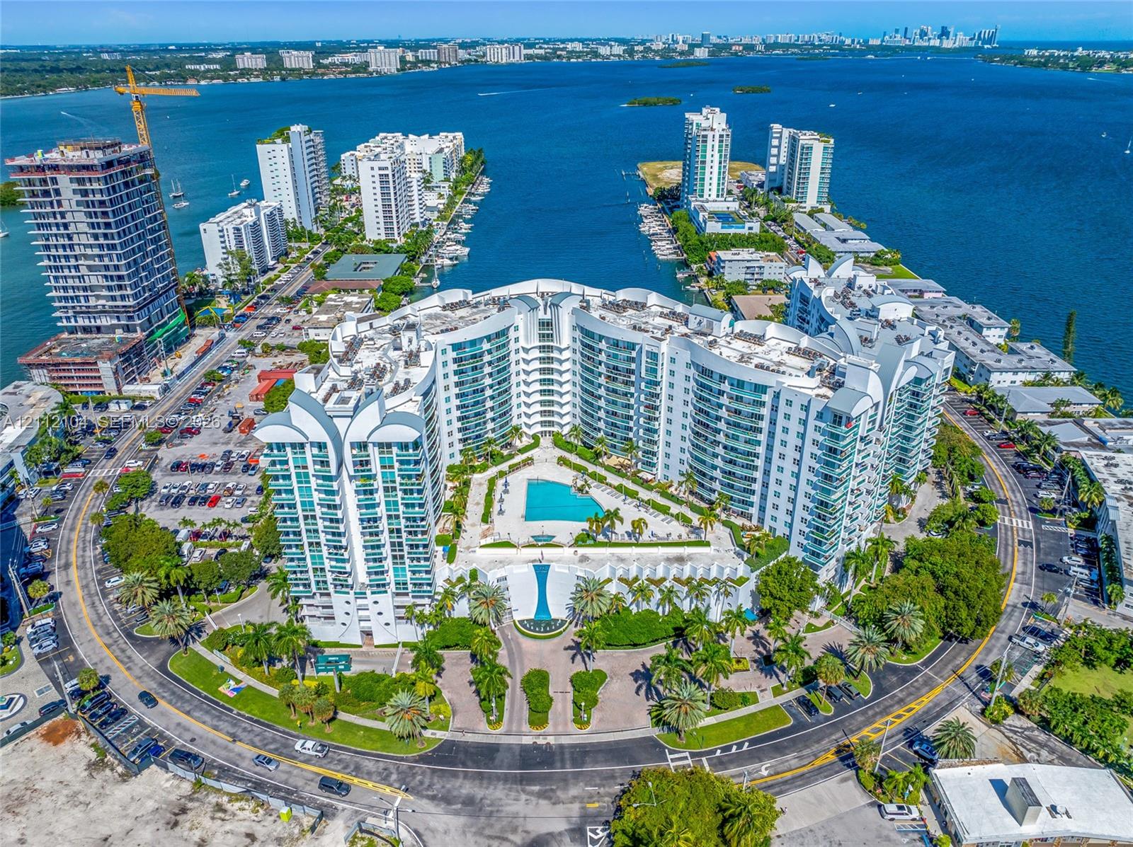 Waterfront residential complex featuring tiered glass-paneled architecture, a central swimming pool with sun deck, landscaped grounds with palm trees, and gated circular drive access