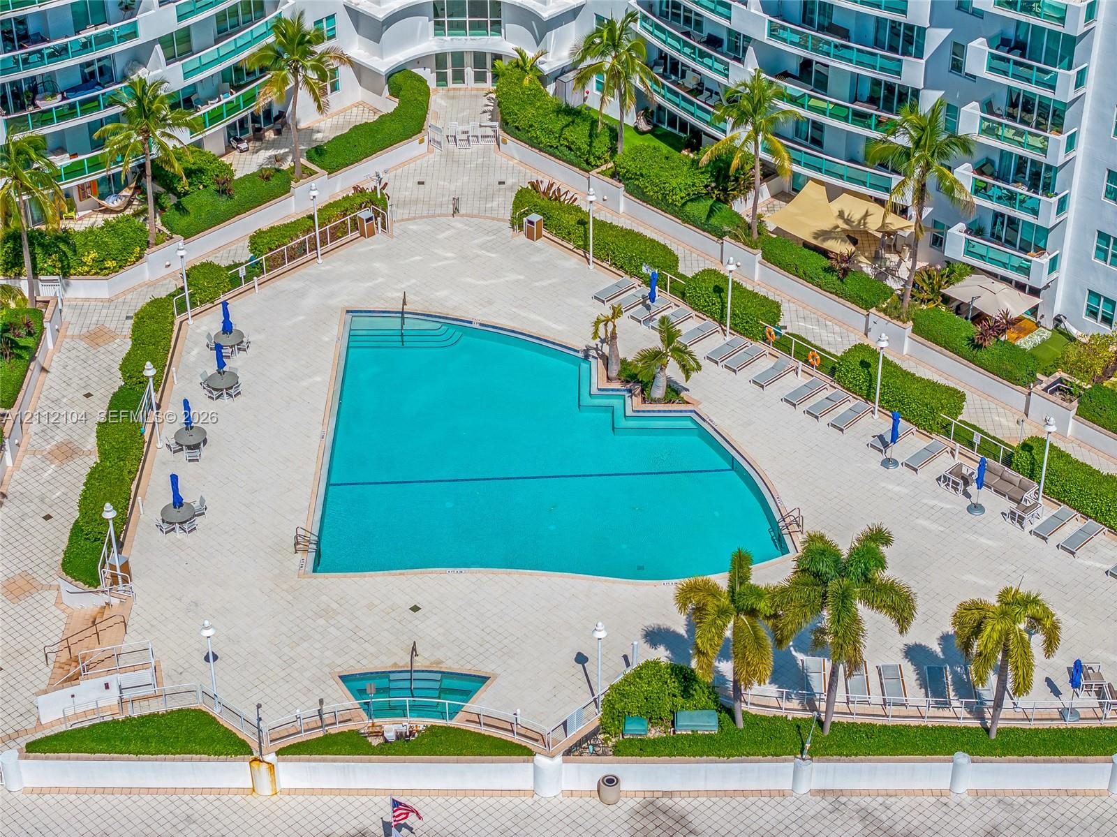 Large swimming pool with tiered entry, paved sun deck, mature palm trees, manicured hedges, and exterior building lighting