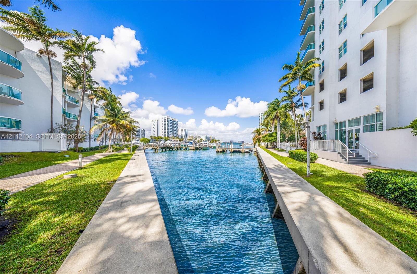 Waterfront canal bulkhead featuring concrete walkways, manicured landscaping, palm trees, and multi-story structures with glass-railing balconies