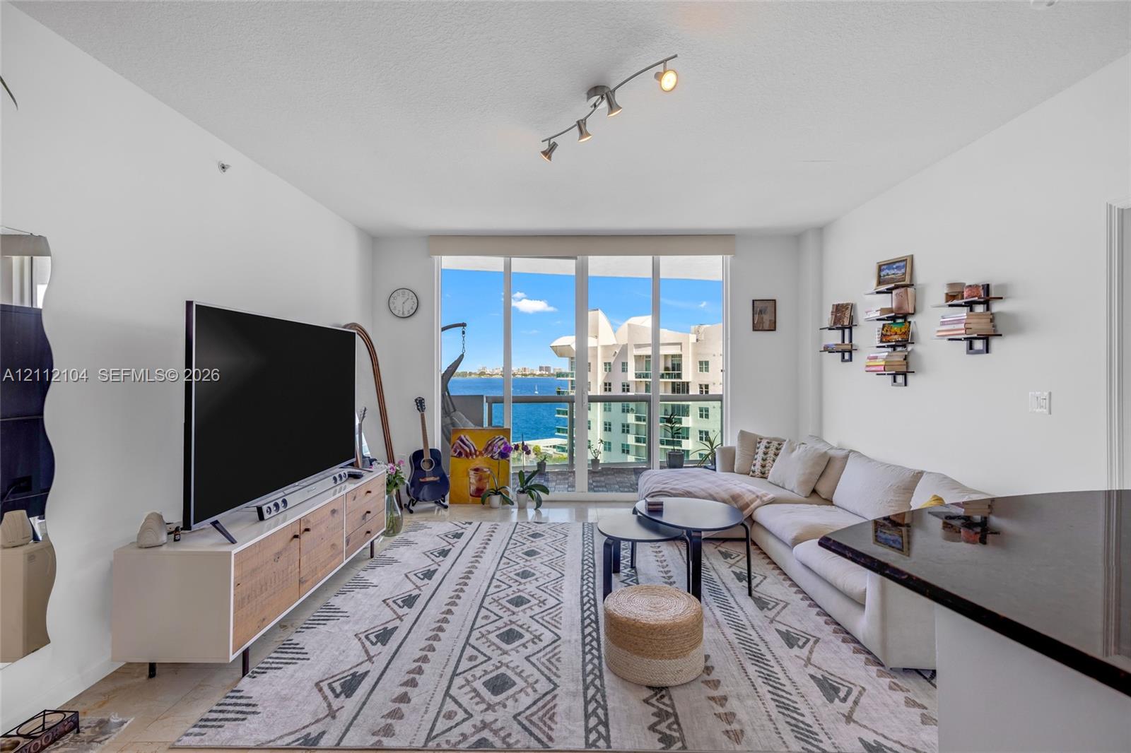 Floor-to-ceiling glass windows, track lighting, and a private balcony featuring a water backdrop