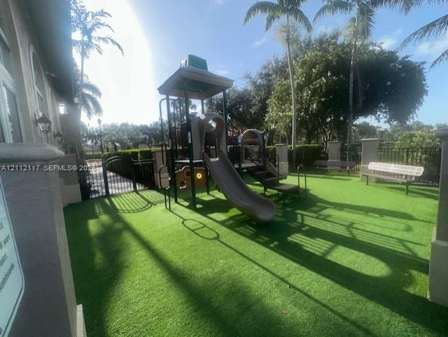 Outdoor recreation area featuring artificial turf, a permanent playground structure with a slide, built-in metal bench seating, and perimeter metal fencing