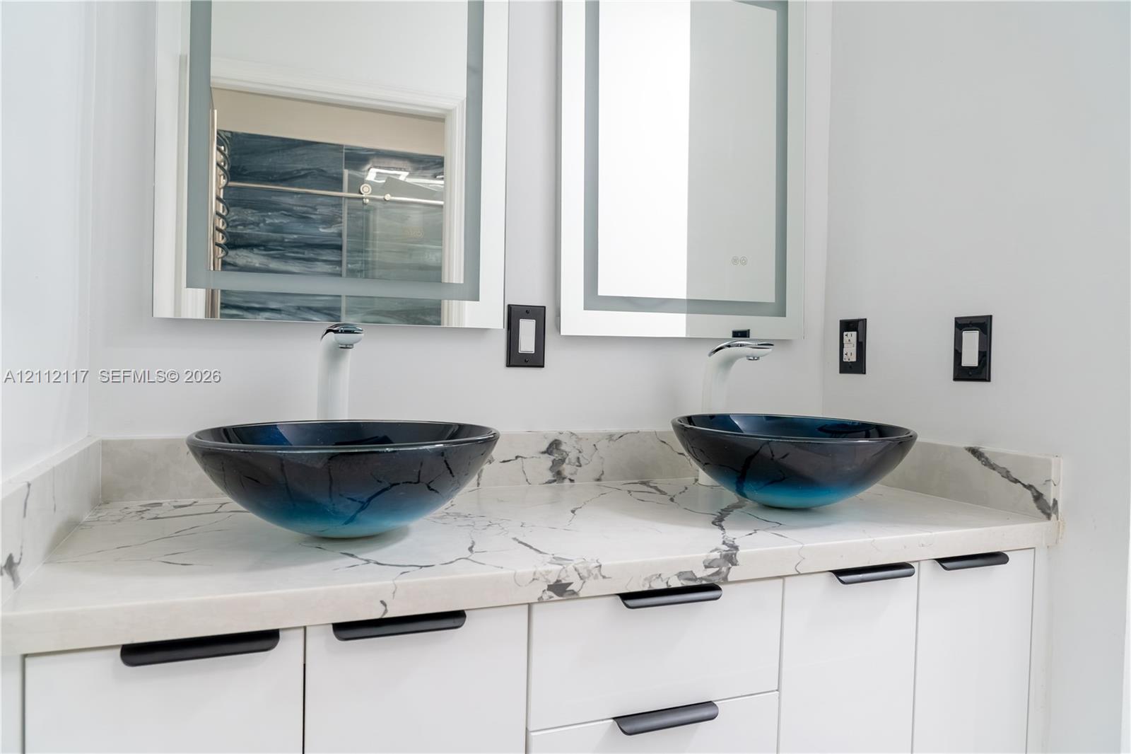 Dual-sink vanity featuring stone-patterned countertops, integrated vessel sinks, white cabinetry with matte black pulls, and illuminated wall-mounted mirrors Dual-sink vanity featuring stone-patterned countertops, integrated vessel sinks, white cabinetry with matte black pulls, and illuminated wall-mounted mirrors
