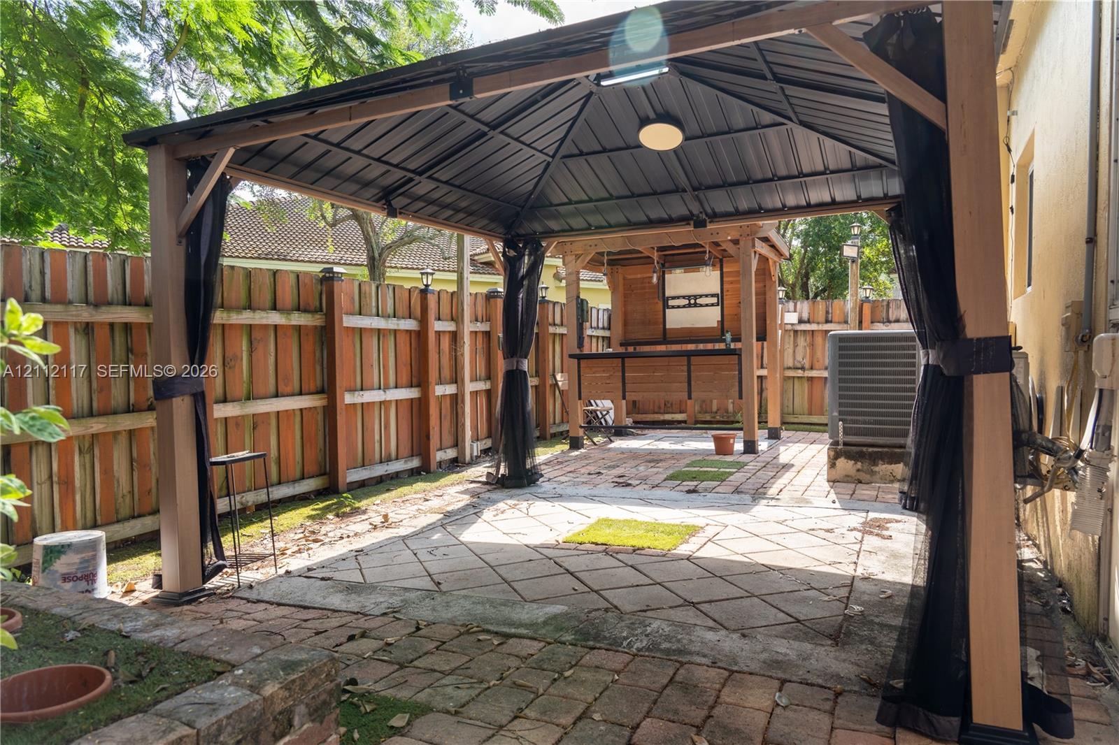 Outdoor patio area featuring a metal-frame gazebo, paved stone surfaces, and wood-panel privacy fencing