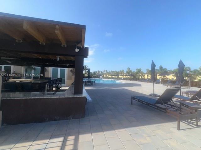 Covered outdoor kitchen featuring a stone-topped bar, wood-beamed ceiling, stone paver patio, and swimming pool