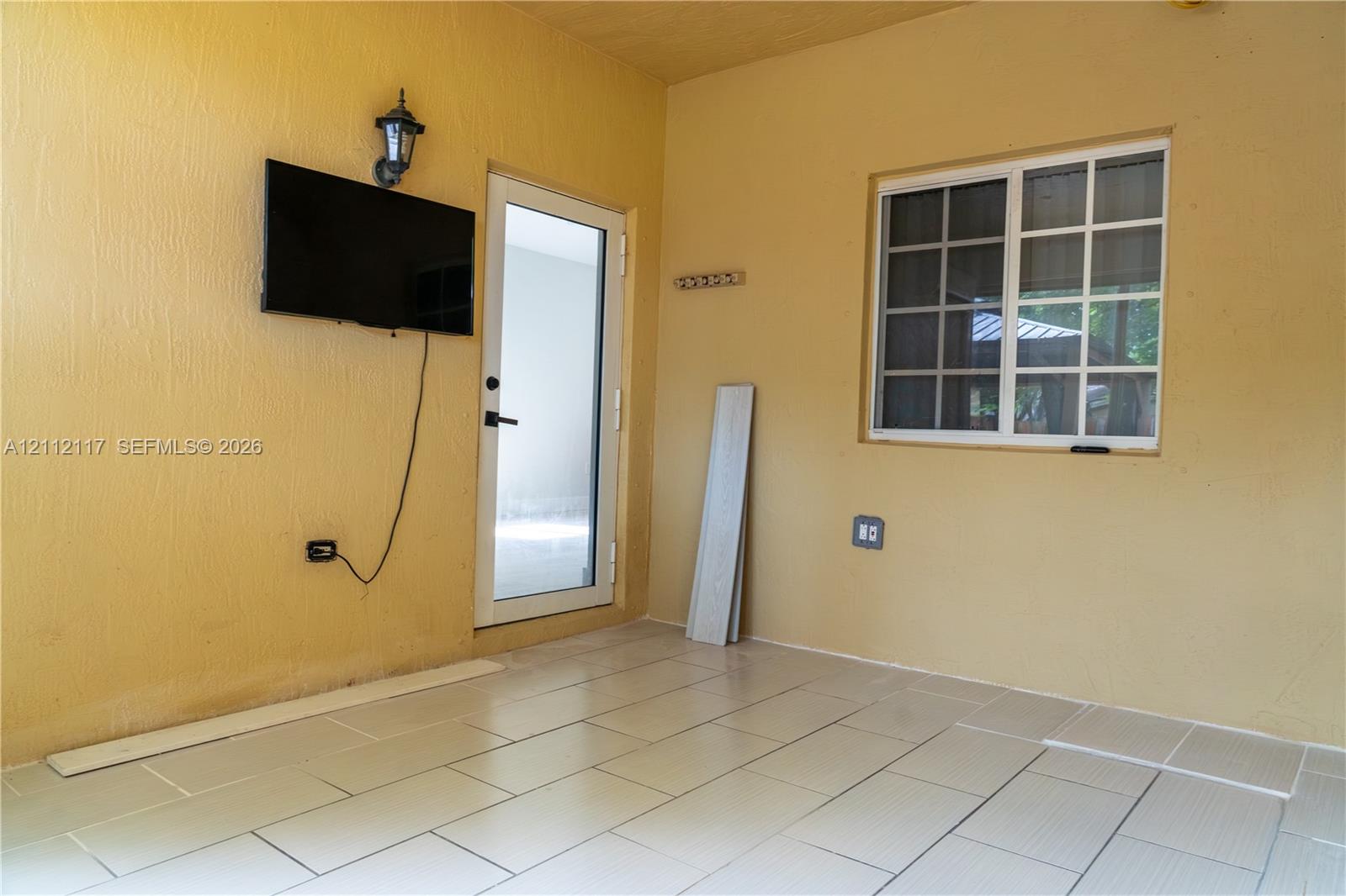 Covered outdoor area featuring neutral-toned tile flooring, a wall-mounted light fixture, and a glass-paneled entry door with a multi-pane window Covered outdoor area featuring neutral-toned tile flooring, a wall-mounted light fixture, and a glass-paneled entry door with a multi-pane window