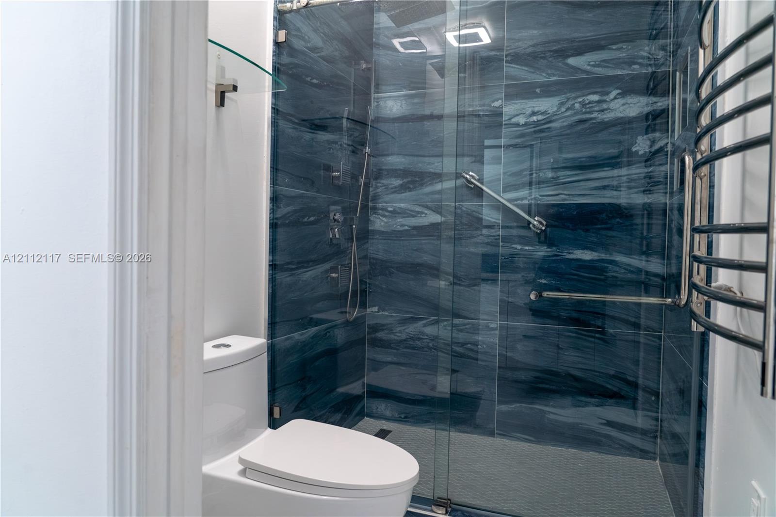 Walk-in shower featuring blue-toned stone-finish tile surround, glass enclosure, wall-mounted shower fixture, and a chrome-finish heated towel rack Walk-in shower featuring blue-toned stone-finish tile surround, glass enclosure, wall-mounted shower fixture, and a chrome-finish heated towel rack