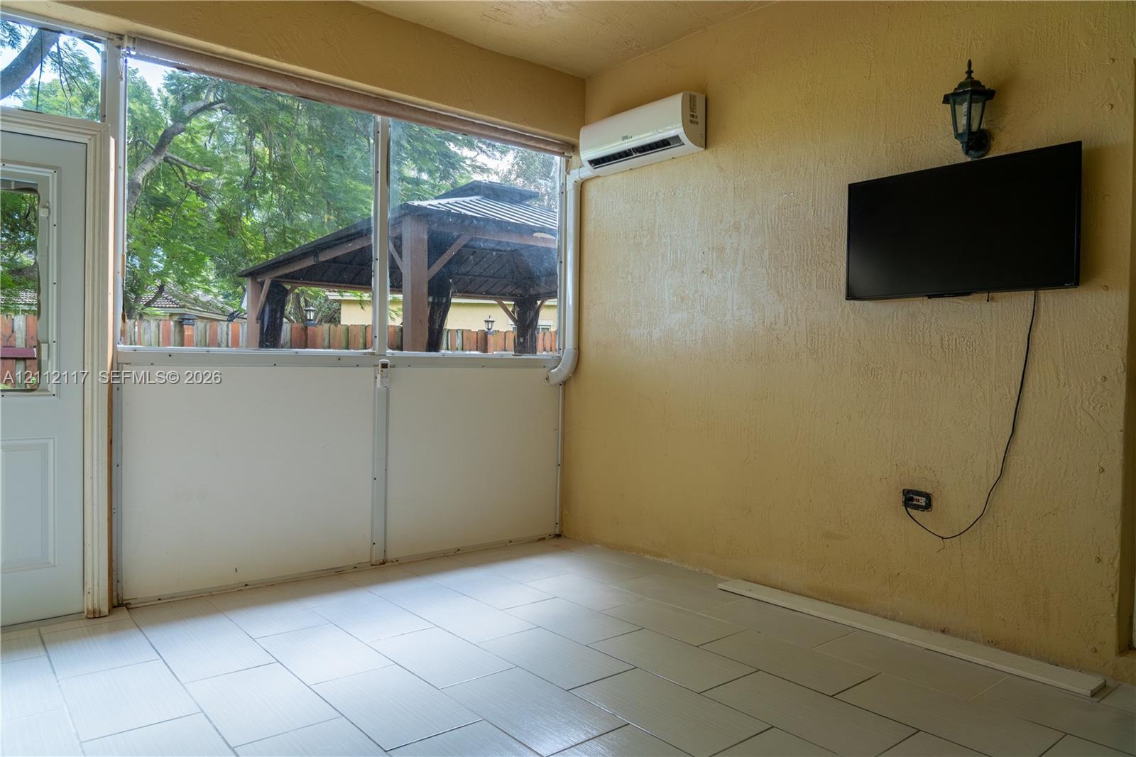 Enclosed patio featuring floor-to-ceiling screen panels, neutral ceramic tile flooring, a wall-mounted ductless air conditioning unit, and an exterior wall-mounted lantern light fixture Enclosed patio featuring floor-to-ceiling screen panels, neutral ceramic tile flooring, a wall-mounted ductless air conditioning unit, and an exterior wall-mounted lantern light fixture
