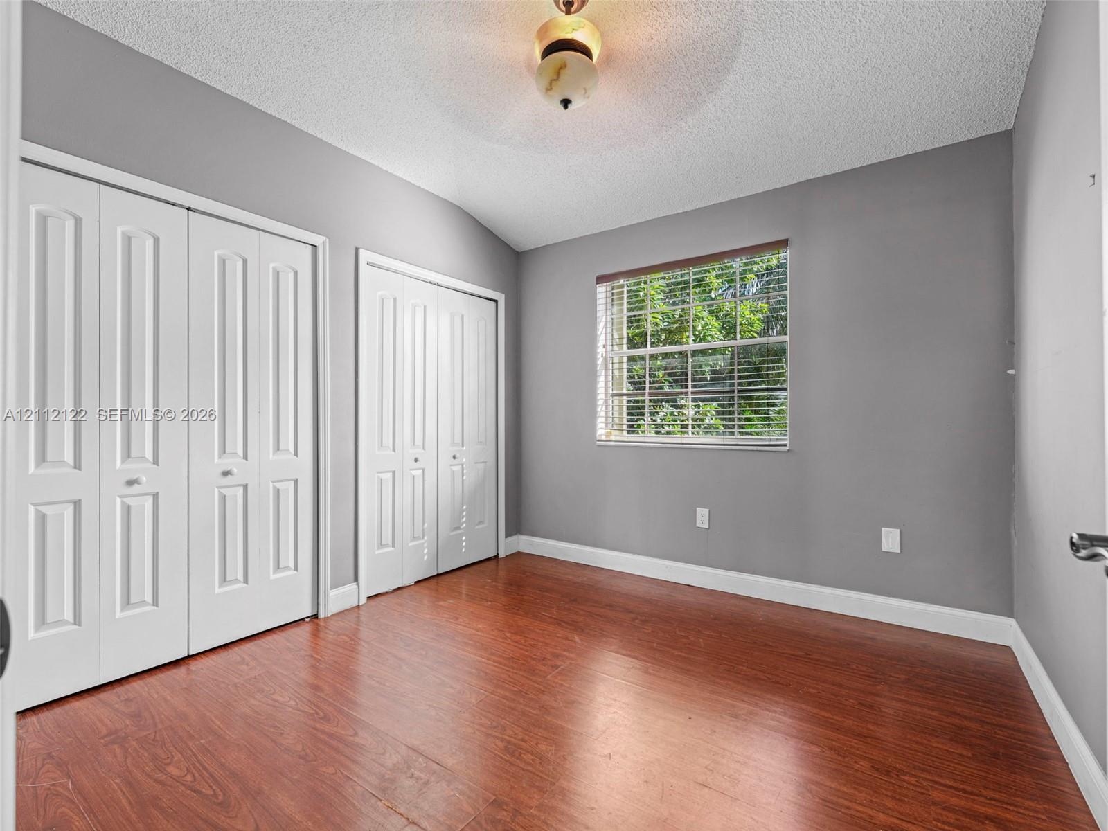 Wood-finish flooring, two sets of bifold closet doors, single-window with horizontal blinds, and a central ceiling-mounted light fixture