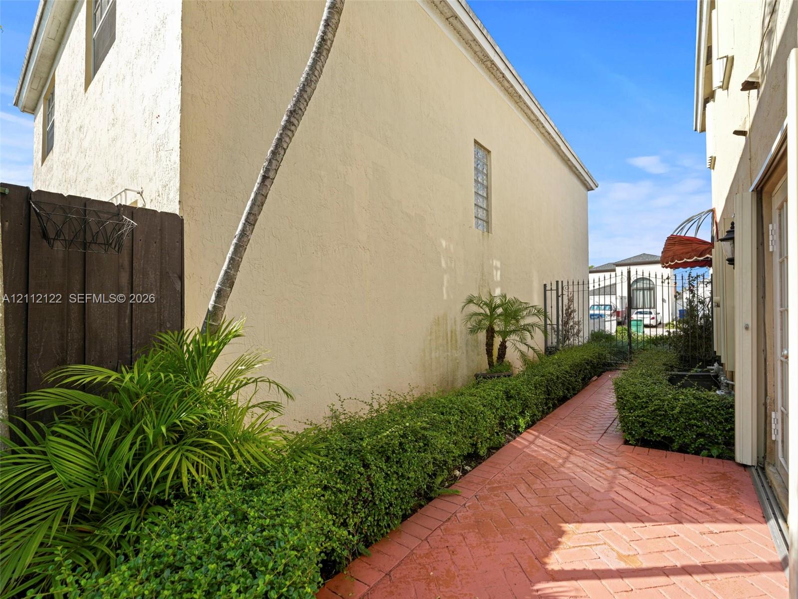 Side exterior featuring herringbone brick paving, mature manicured shrubbery, stucco siding, and a vertical glass block accent window