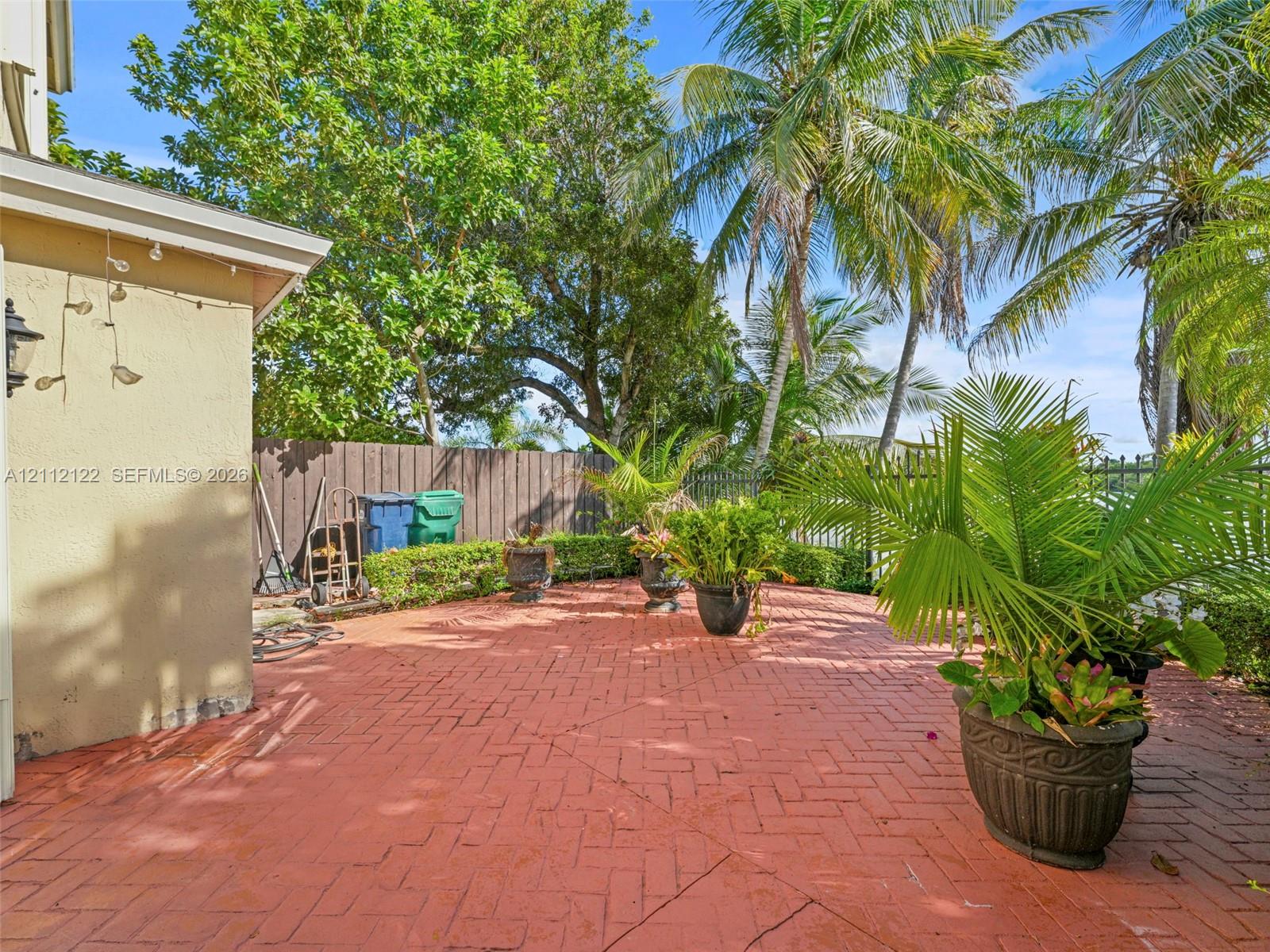 Herringbone-patterned brick patio featuring a perimeter hedge, mature palm trees, and metal fencing