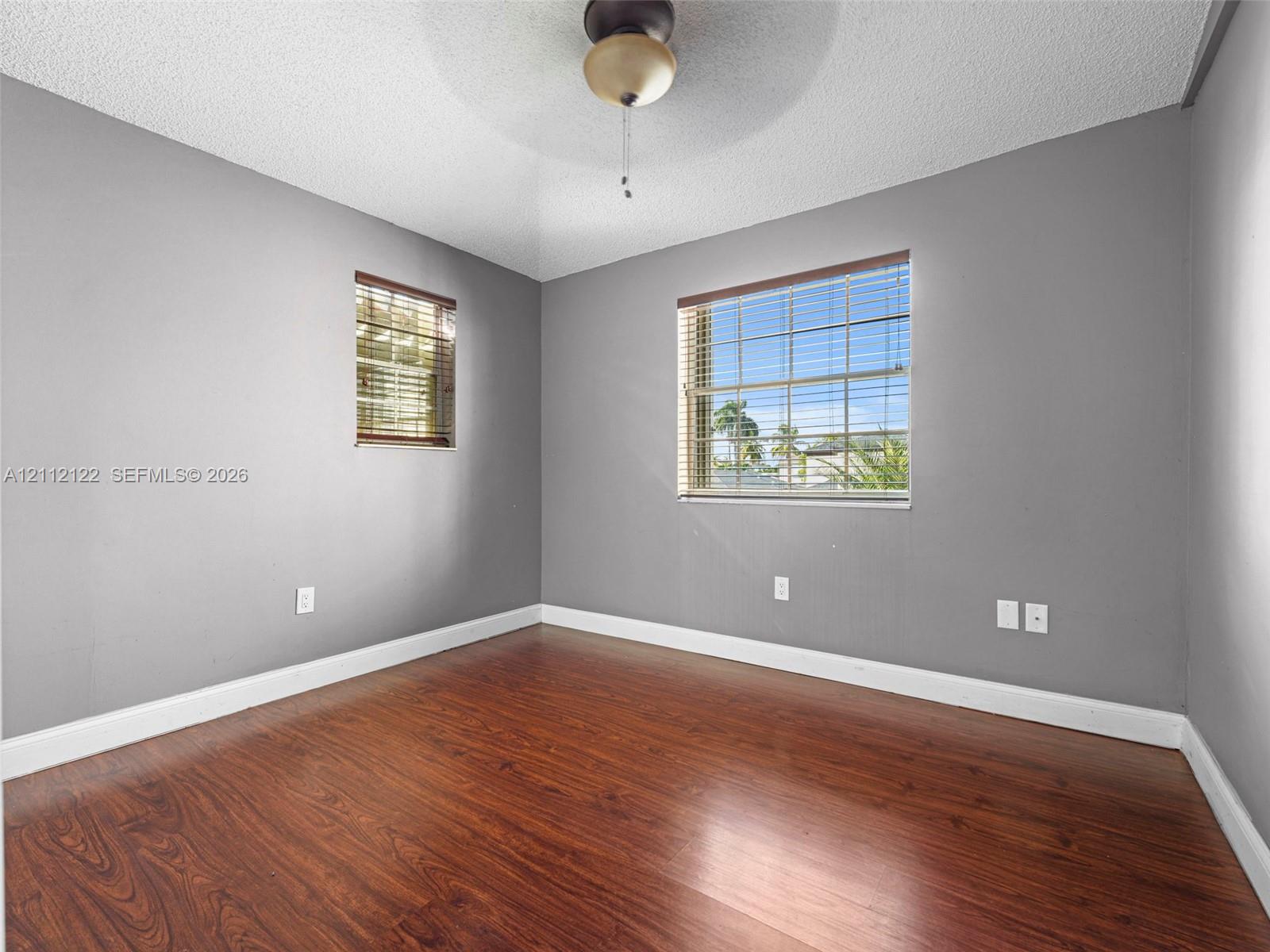 Wood-finish flooring, neutral-toned walls, ceiling-mounted light fixture with fan, and dual windows with horizontal blinds