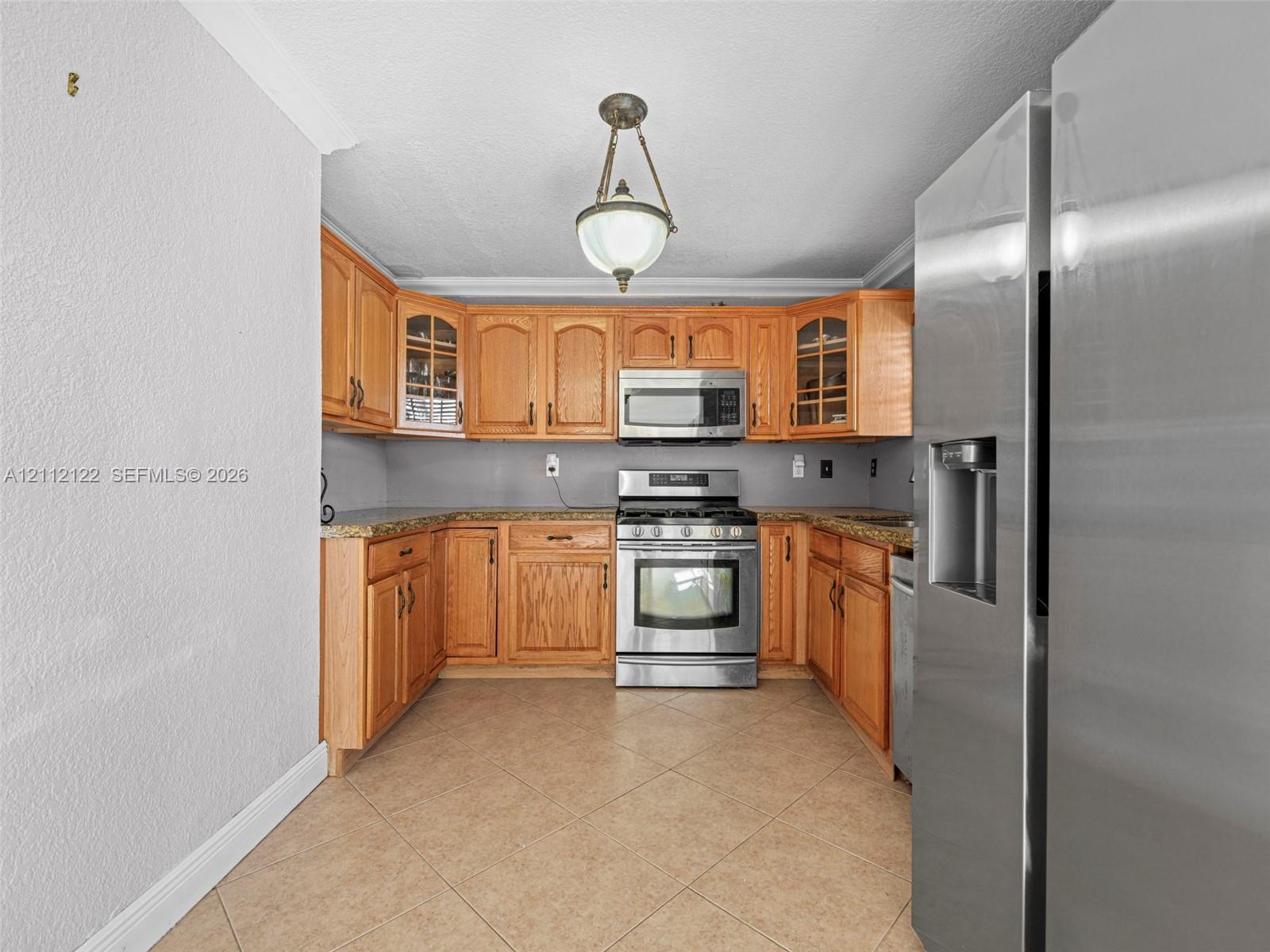 Kitchen featuring wood cabinetry, stone-surface countertops, tile flooring, stainless steel appliances, and a central pendant light fixture
