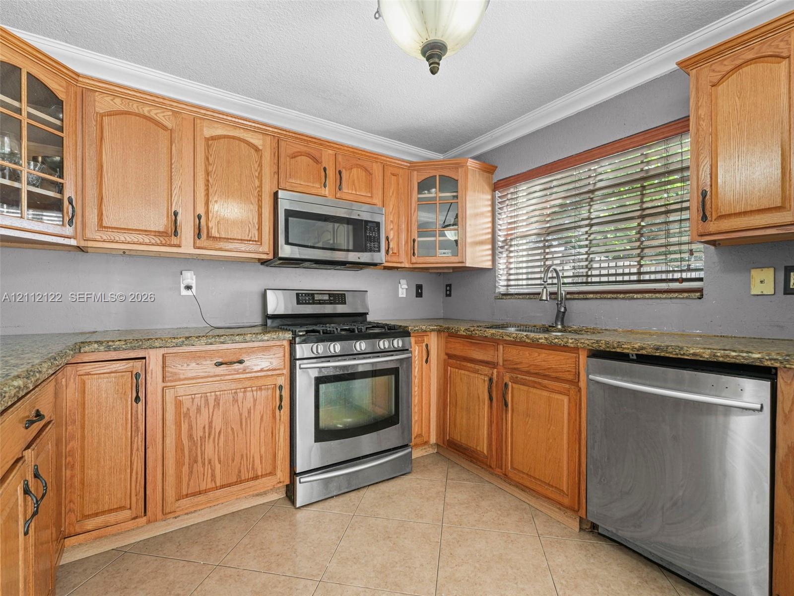Kitchen featuring wood cabinetry, stone-finish countertops, stainless steel appliances, ceramic tile flooring, and a window with blinds