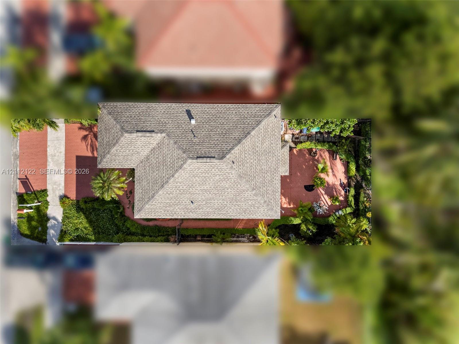 Aerial view featuring a multi-faceted shingle roof, concrete driveway, and paved patio area surrounded by mature landscaping