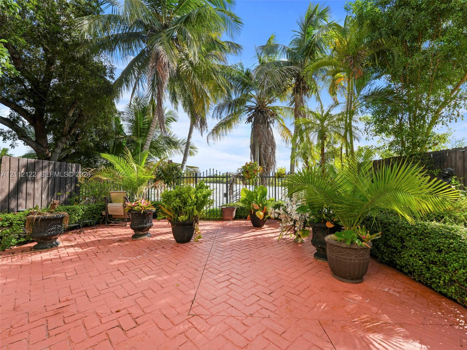 Herringbone-patterned brick patio featuring a perimeter of sculpted hedging, mature palm trees, and decorative metal perimeter fencing