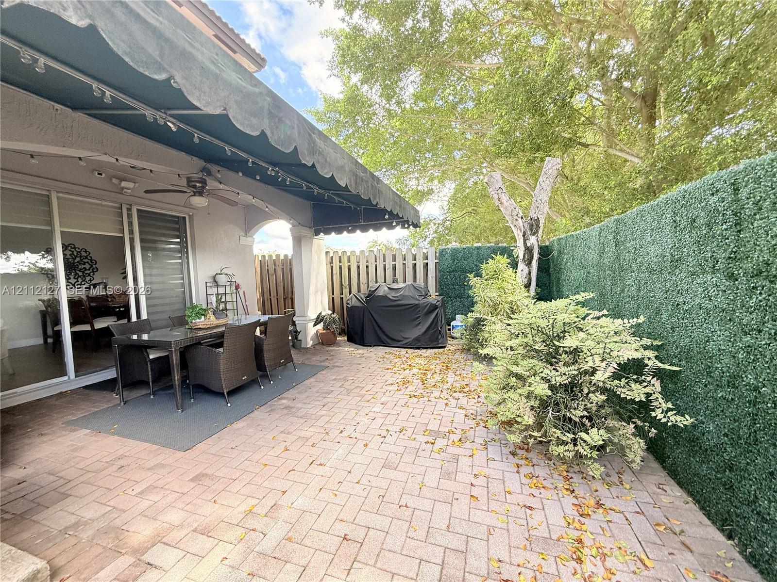 Covered patio featuring a herringbone-patterned brick floor, retractable fabric awning, arched structural columns, and a privacy hedge