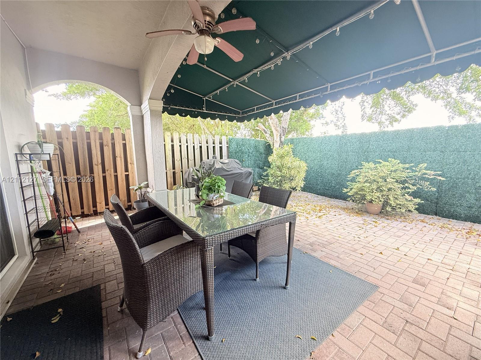 Covered brick patio featuring a green retractable awning, architectural archway, and ceiling fan, enclosed by wood and privacy-screening fencing
