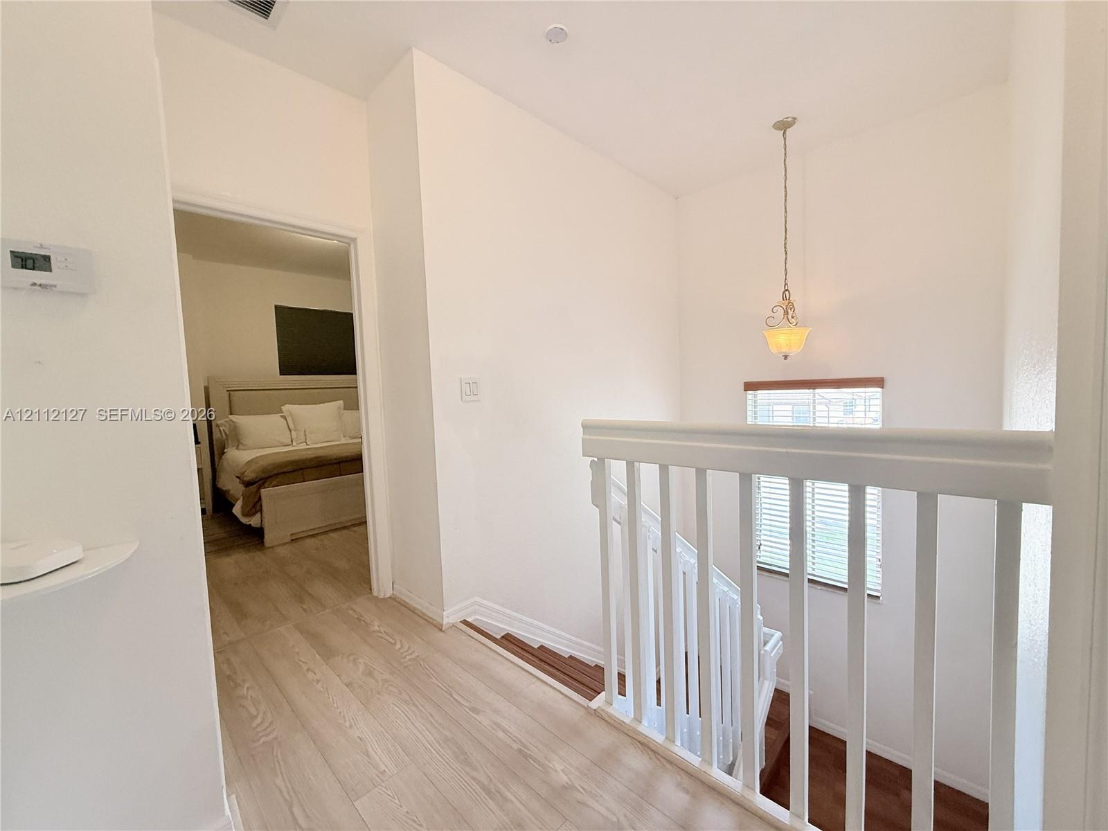 Hallway featuring wood-finish flooring, white spindle railing, a pendant light fixture, and horizontal window blinds