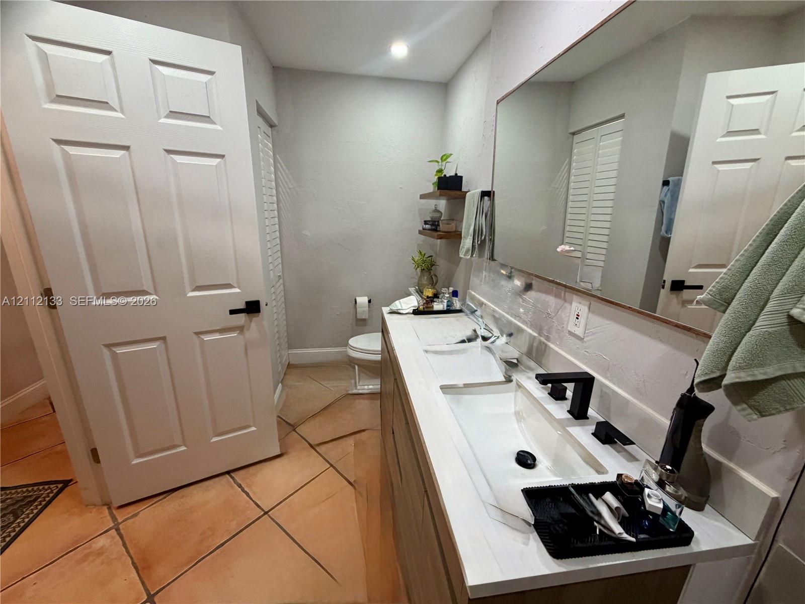 Dual-sink vanity with matte black fixtures, stone-surface countertop, full-width mirror, and terracotta tile flooring