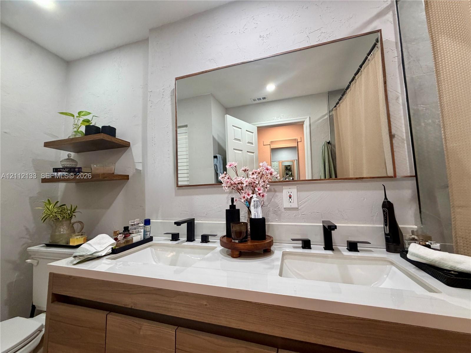 Dual-sink vanity with wood-finish cabinetry, white countertop, matte black widespread faucets, and two built-in wood-finish floating shelves