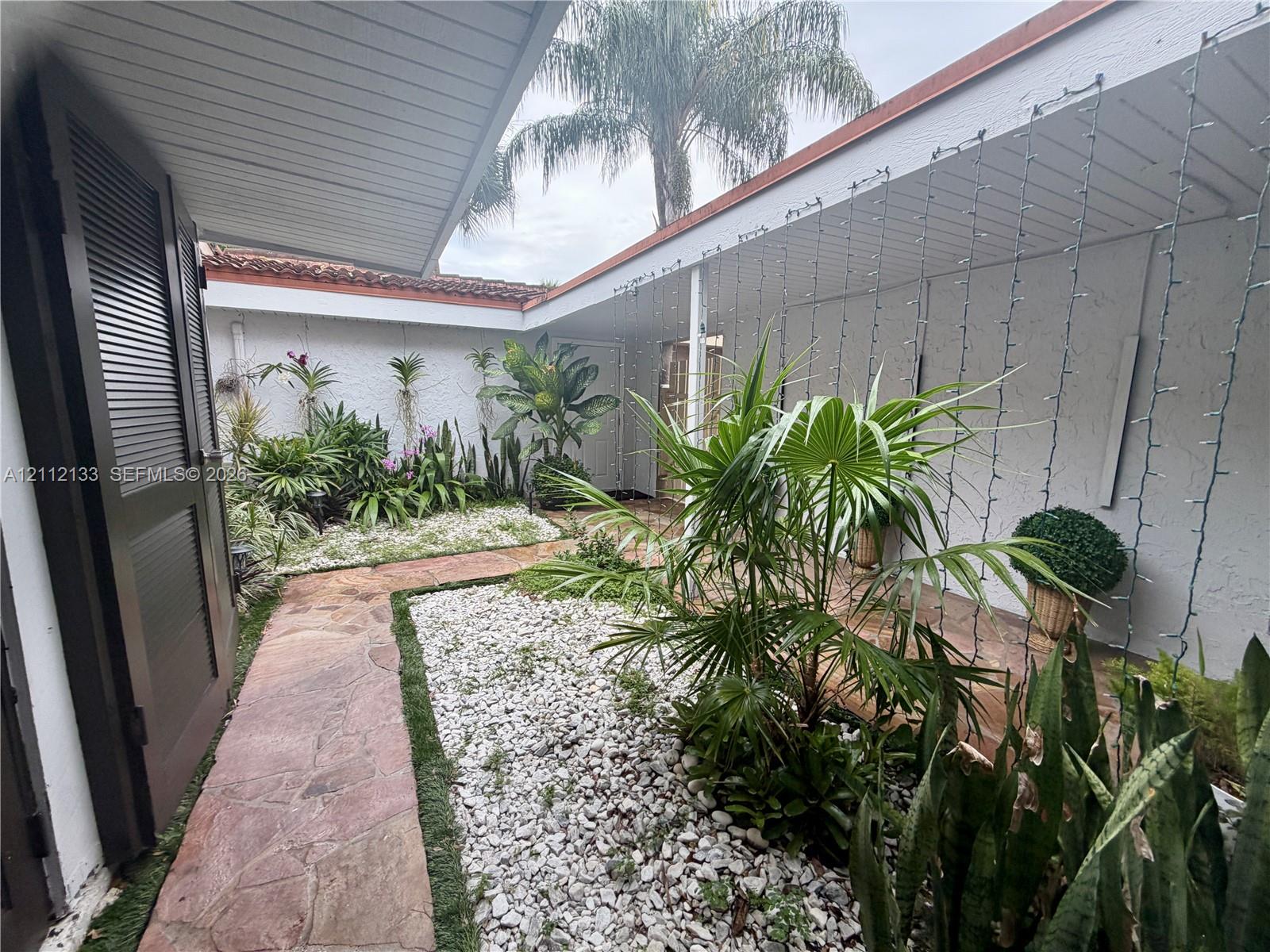 Enclosed courtyard featuring flagstone pathways, white stone ground cover, mature tropical plantings, and a covered walkway