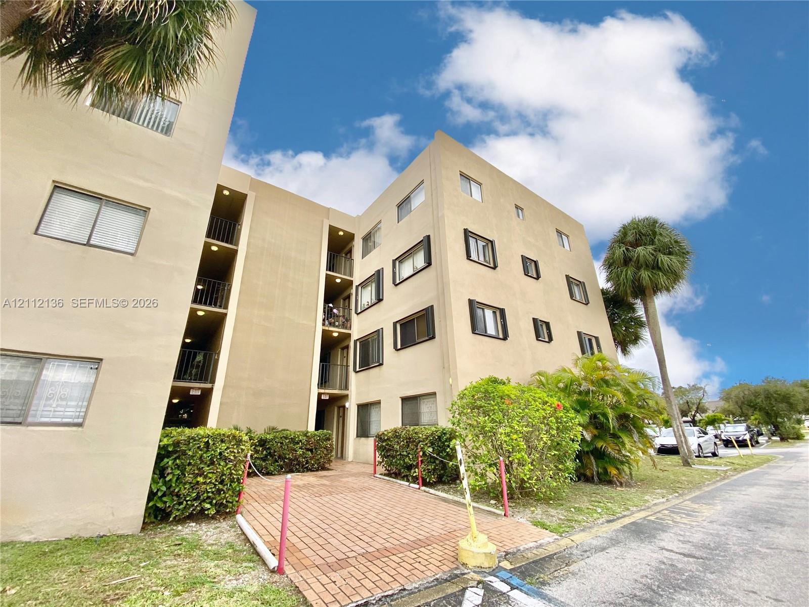 Multi-story exterior featuring tan stucco facade, brick-paved walkway, and mature landscaping