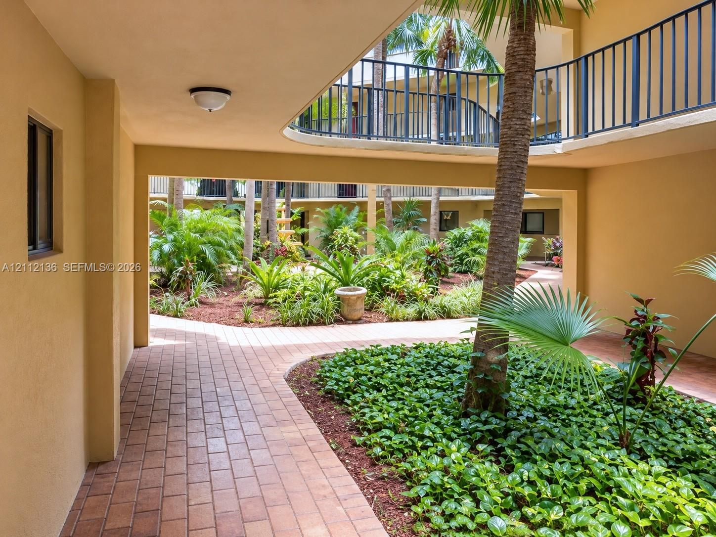 Open-air courtyard featuring brick paver walkways, lush tropical landscaping, mature palm trees, and metal railed balconies