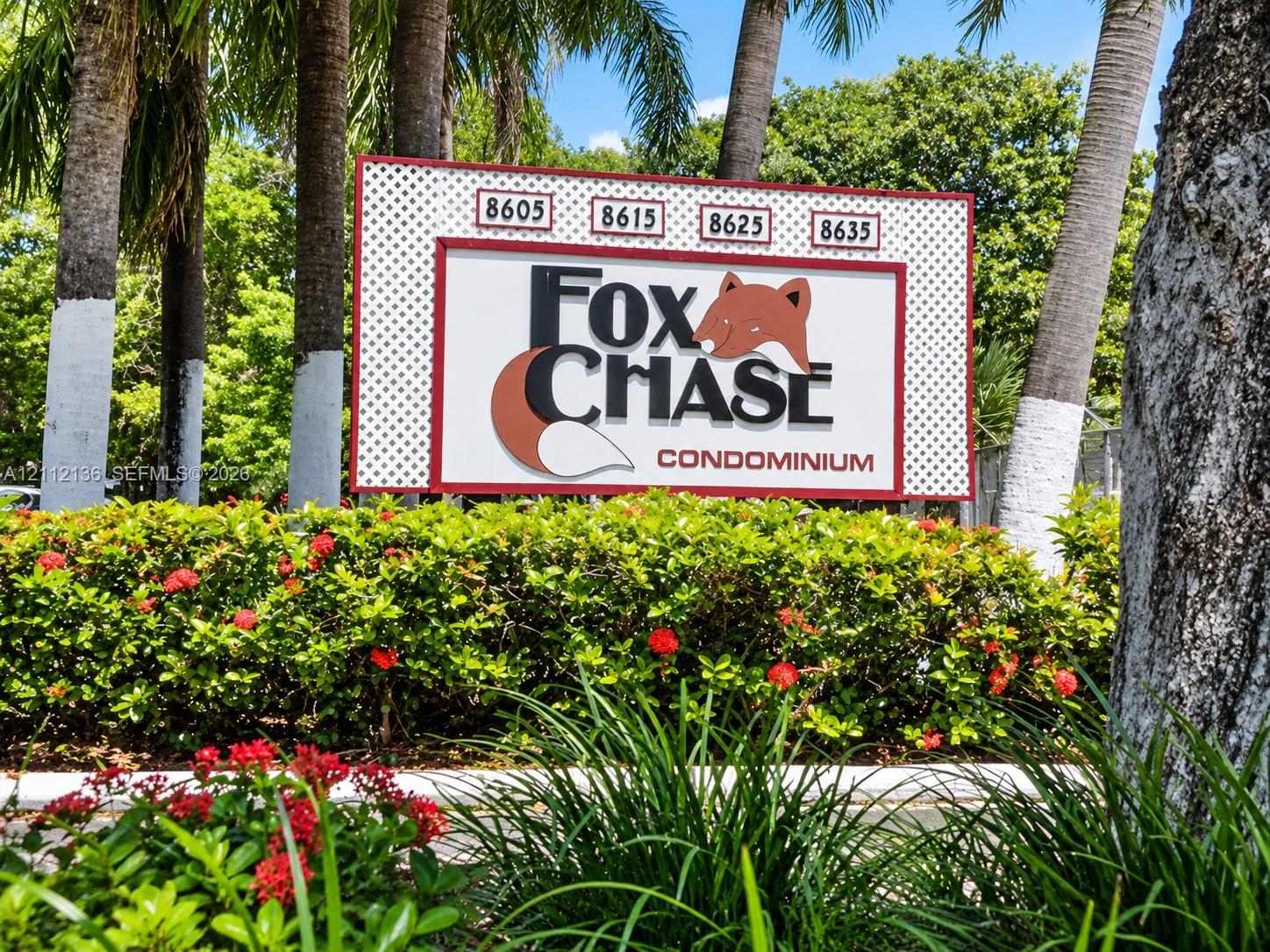 Community entrance signage for Fox Chase Condominium featuring mature palm trees and manicured shrubbery with flowering plants Community entrance signage for Fox Chase Condominium featuring mature palm trees and manicured shrubbery with flowering plants