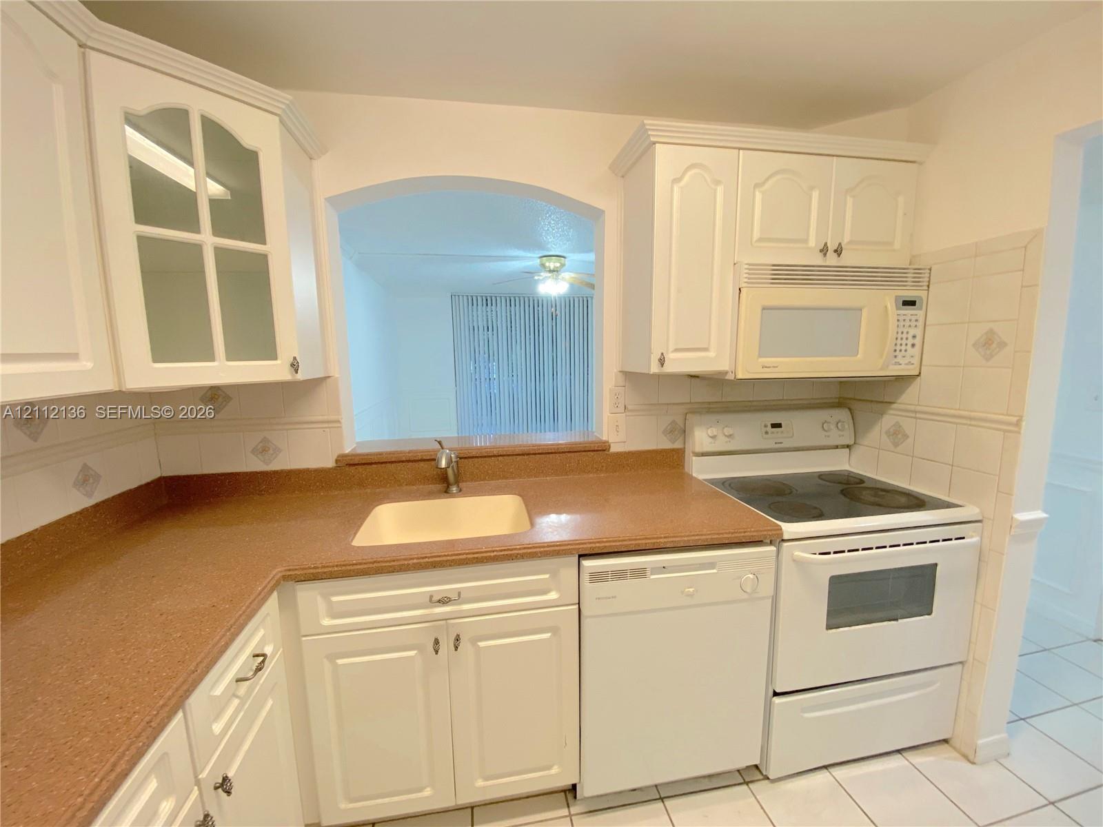Kitchen featuring white cabinetry with glass-panel upper doors, stone-composite countertops, tile backsplash, built-in appliances, and a pass-through window