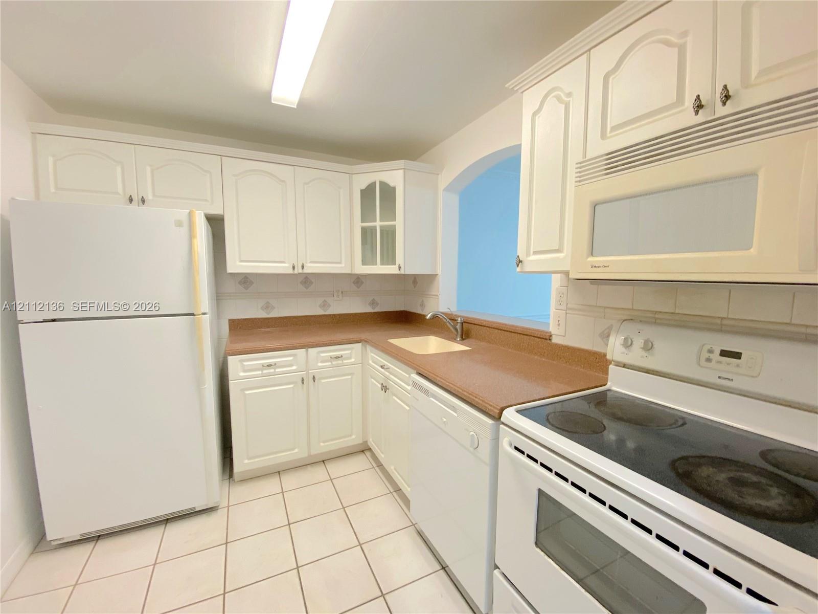 Kitchen featuring white cabinetry with glass-panel accents, solid-surface countertops, tile flooring, and integrated white appliances