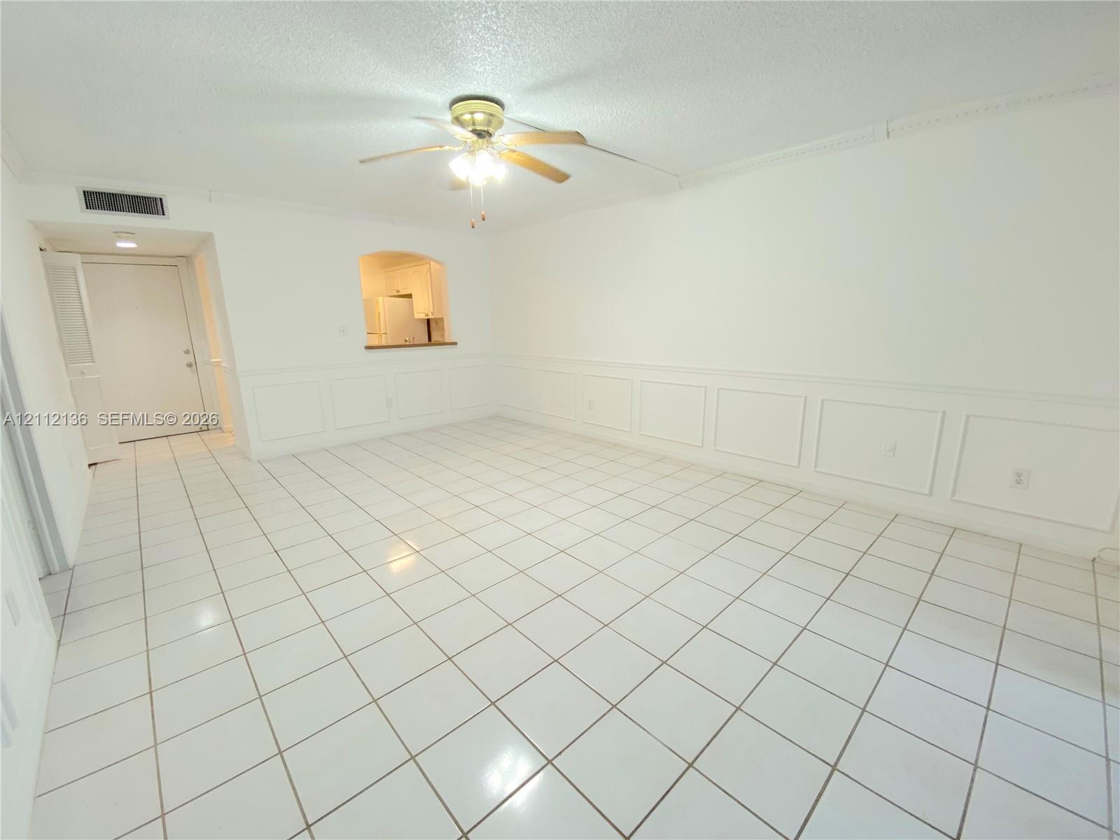 Neutral-toned interior space featuring light-colored square tile flooring, decorative wainscoting, an arched pass-through window, and a ceiling-mounted fan with integrated lighting