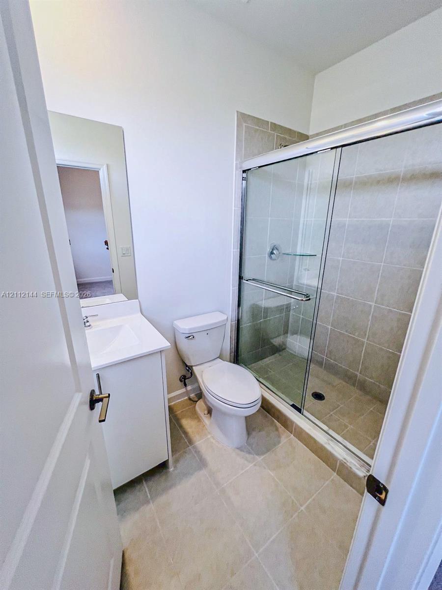 Bathroom featuring a single-sink vanity with a solid-surface countertop, tiled walk-in shower with sliding glass doors, and neutral ceramic tile flooring