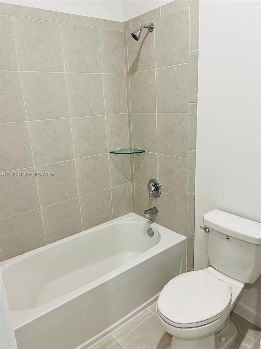 Bathroom featuring a porcelain bathtub, integrated shower fixture, neutral-toned wall tile surround, glass corner shelf, and standard-height toilet