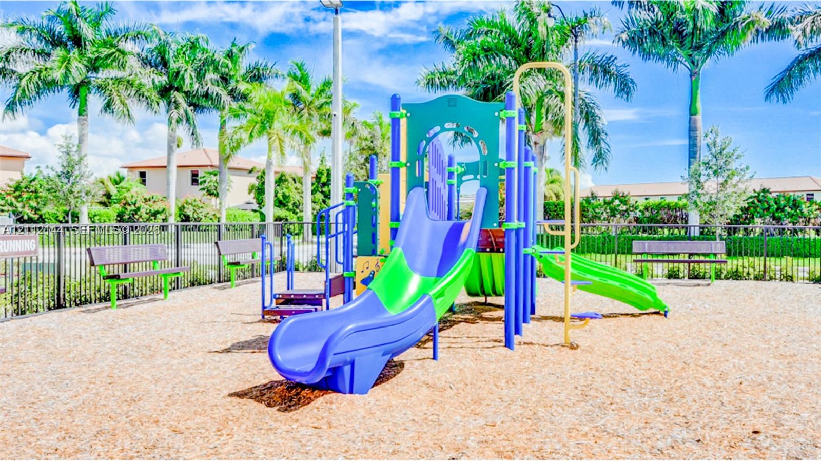 Recreational area featuring a multi-component play structure with dual slides, wood-chip ground cover, and metal bench seating enclosed by perimeter fencing